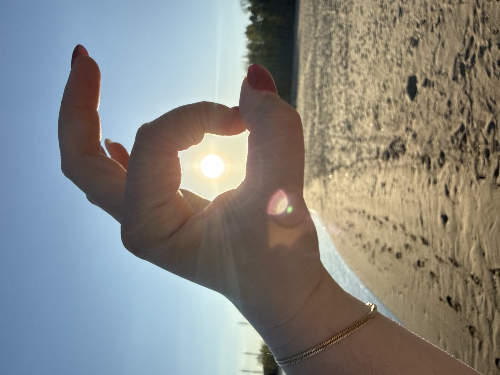 A person's right hand, forming an "OK" sign with the sun visible through the circle, is the main focus of the image. The hand has a gold-colored bracelet on the wrist and red-painted fingernails. The background consists of a beach, with possible footprints visible in the sand, and a blue sky. The location is the city of Noviy, Russia.