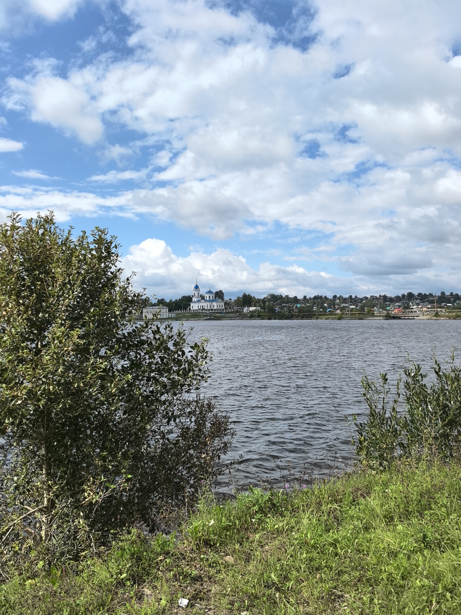 The image depicts a wide, calm body of water, likely the Kama River, in the city of Dobryanka, Russia. On the far bank, a small cluster of buildings is visible, including a prominent white church with blue domes. The sky is partly cloudy, with large white clouds against a blue backdrop. The foreground is dominated by lush green grass and foliage, with a large tree on the left side partially obscuring the view of the water and the distant settlement. The water's surface is rippled, suggesting a gentle breeze. The scene is a natural landscape with a man-made structure integrated into it.