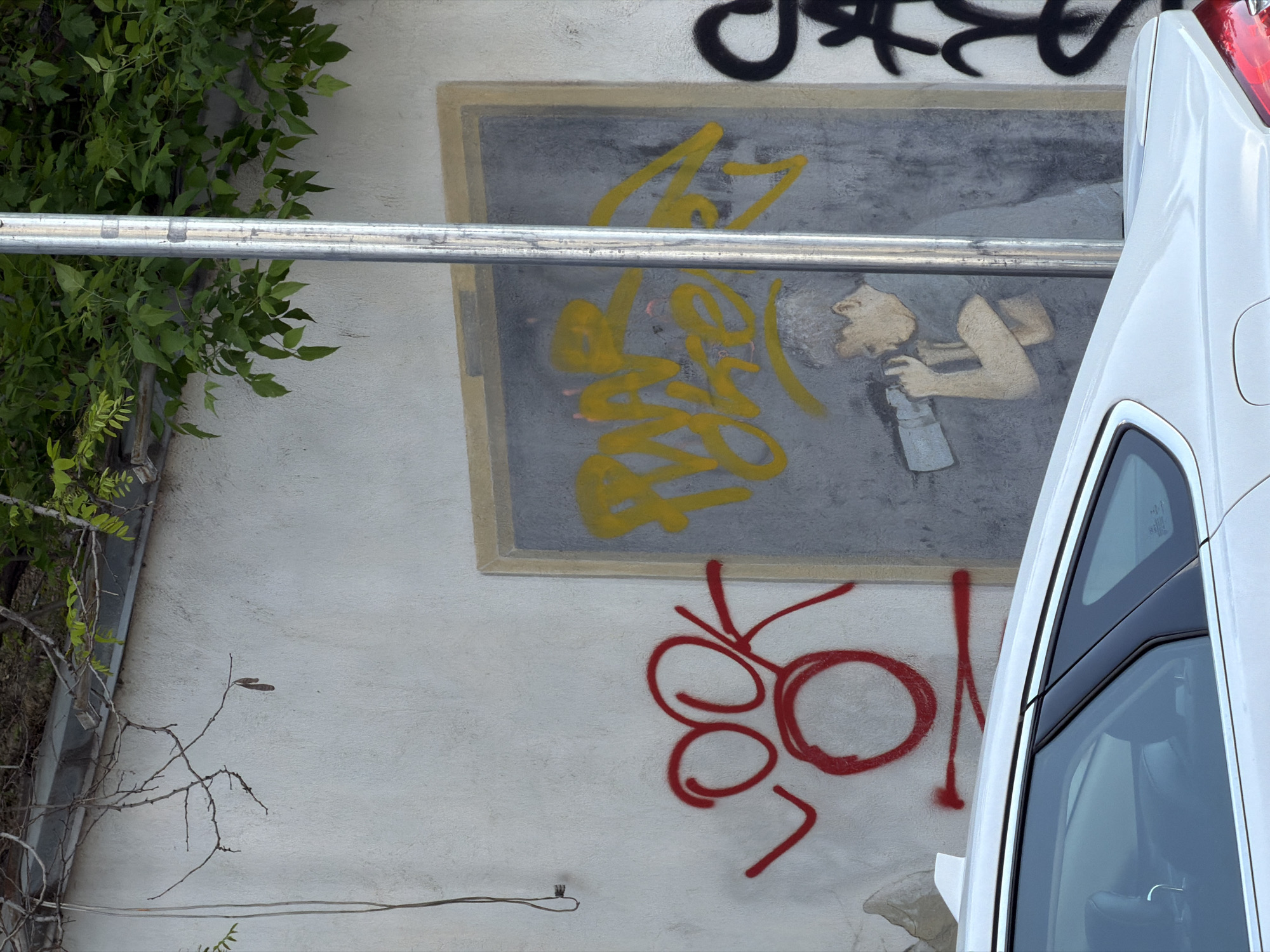 This image depicts a section of a light-colored, textured urban wall, densely covered with various forms of street art and graffiti. The scene is presented from a side-on perspective, partially obscured by foreground elements.
Centered on the wall is a rectangular mural with a grey background, framed by a light brown border. Within this mural, a beige-toned illustration portrays a person, possibly male, with short hair or a beard, holding a silver spray can as if actively painting or inhaling. Overlaid on this mural are vibrant yellow graffiti tags, consisting of abstract, flowing letters and shapes. Above the mural, partially visible, are black graffiti elements, appearing as stylized lettering. Below the mural, red graffiti features prominent circular shapes and lines, potentially spelling "LOO" or "LOOK," though the last part is ambiguous.
A horizontal silver metal bar, possibly a scaffolding pole or railing, traverses the middle of the image, partially obscuring the yellow graffiti and the painted figure. On the right, the white bodywork and windows of a vehicle, likely a van or SUV, are visible, blocking a significant portion of the wall and some of the red graffiti. A red taillight is discernible at the top right. To the left, lush green foliage and several bare branches of a plant extend into the frame, providing a natural element against the concrete wall.
The lighting suggests daytime, possibly under an overcast sky or in shadow, as there are no strong directional shadows. The setting is an urban environment, reflecting a dynamic display of street art in Tbilisi, Georgia.