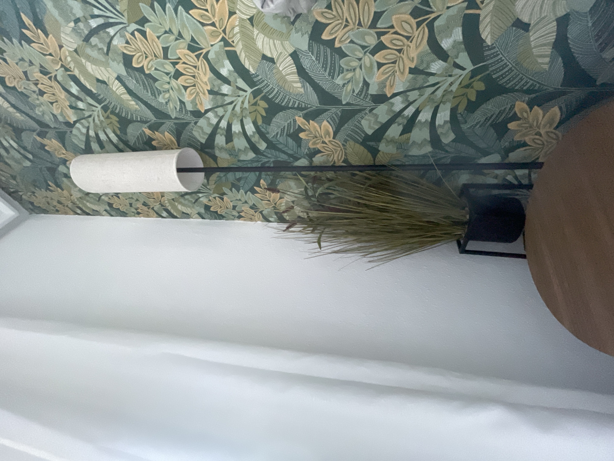 The image shows a section of a room, likely in a residential setting. A white, cylindrical lamp with a black supporting arm is mounted on a wall. The wall behind the lamp is covered with a green and tan botanical wallpaper. A decorative plant with long, thin leaves sits in a black pot. To the right of the plant, a round, wooden table is partially visible. The lower portion of the image is occupied by a white curtain or drapery. The location, Rojales, Spain, is not discernible from the image alone.