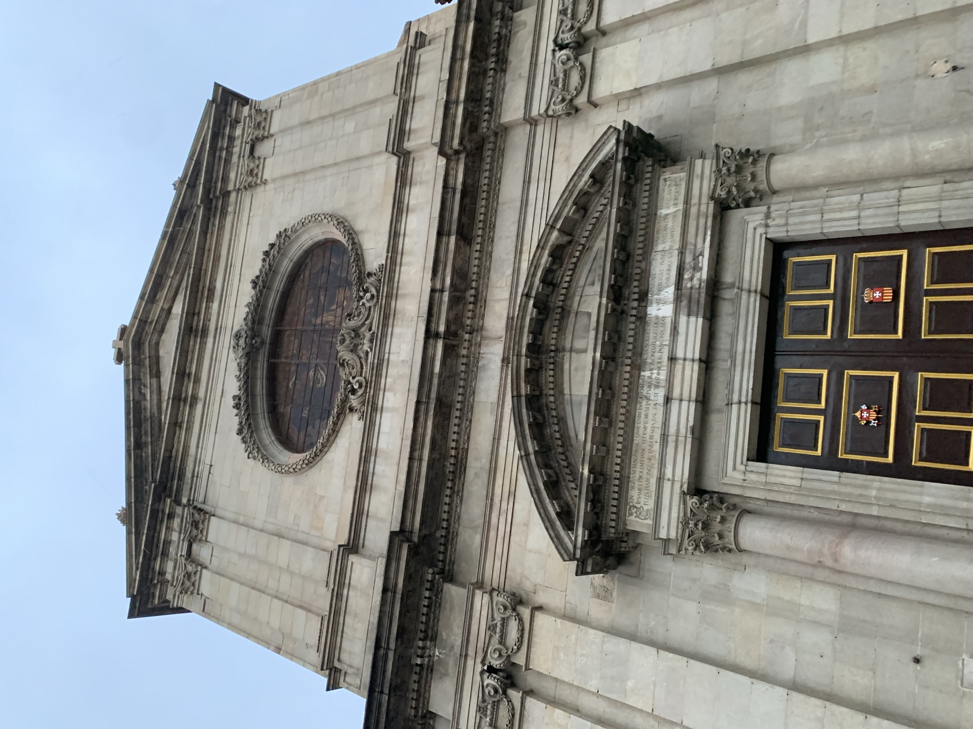 This is a close-up of the facade of the Basilica of Saints Justus and Pastor in Barcelona, Spain. The architecture is light-colored stone, featuring a large oval window, a rounded architectural detail with text, and a doorway with a dark door trimmed in gold. There is a coat of arms on the door. The photo is taken from a low angle, looking up at the building. The sky is visible in the top corners.