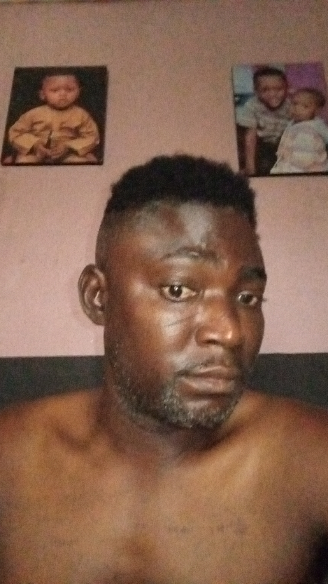 A man with dark skin, a trimmed beard, and a closely cut hairstyle is centered in the frame. He appears to be shirtless, with his shoulders visible. Behind him on the wall are two framed pictures. The left picture depicts a baby in a yellow garment. The right picture shows two children, one seemingly older than the other, with the younger child wearing a light-colored dress. The wall is a light pink color. The location is unknown.