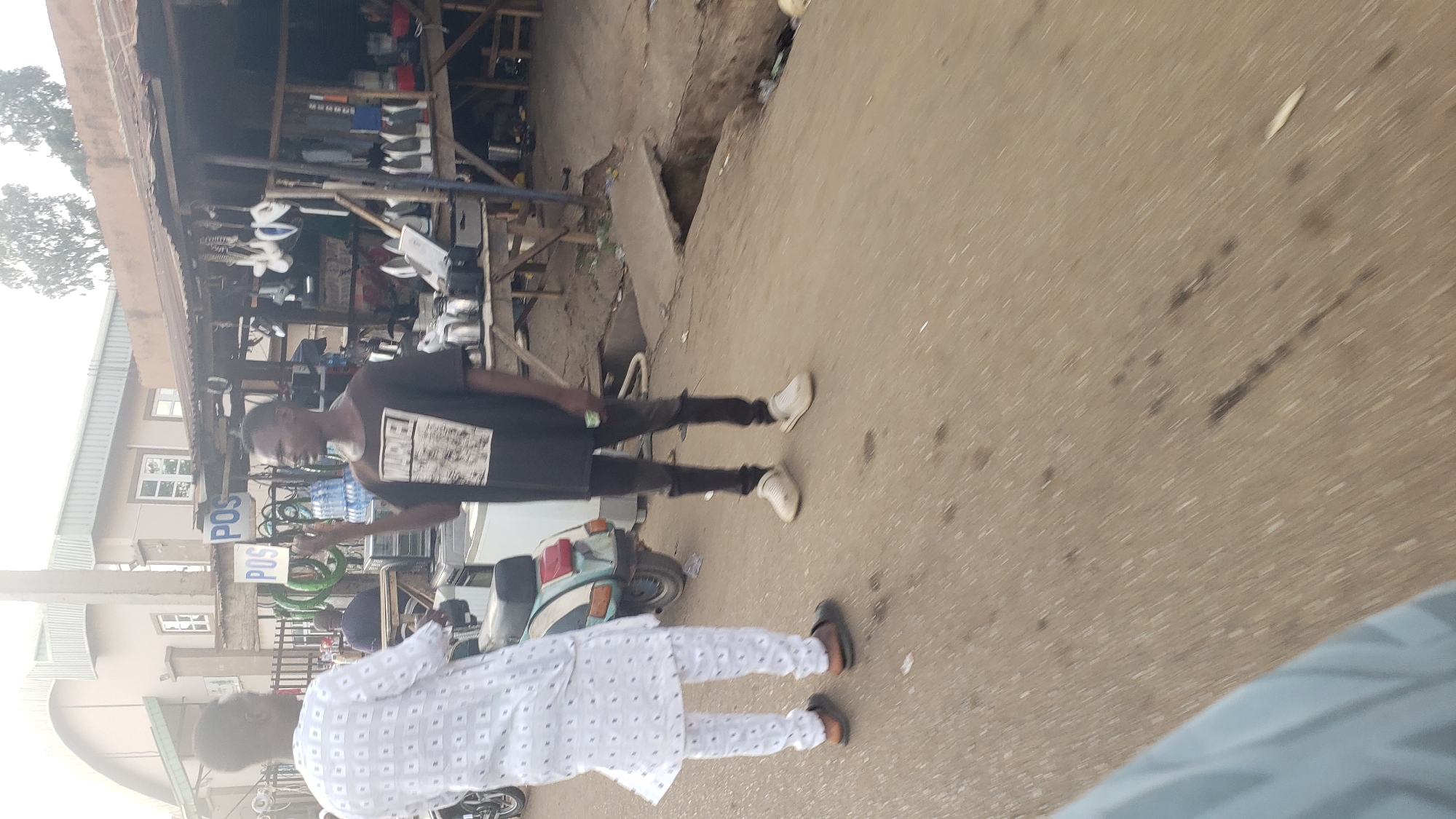 The image depicts a street scene in Kawo, Nigeria. The foreground shows two men standing on a paved surface. One man is wearing a white patterned garment while the other is in a dark t-shirt and dark trousers. A motorcycle is parked between them. Behind the men is a shop selling various metal items. A POS sign is also visible near the shop. A building with windows is present in the background.