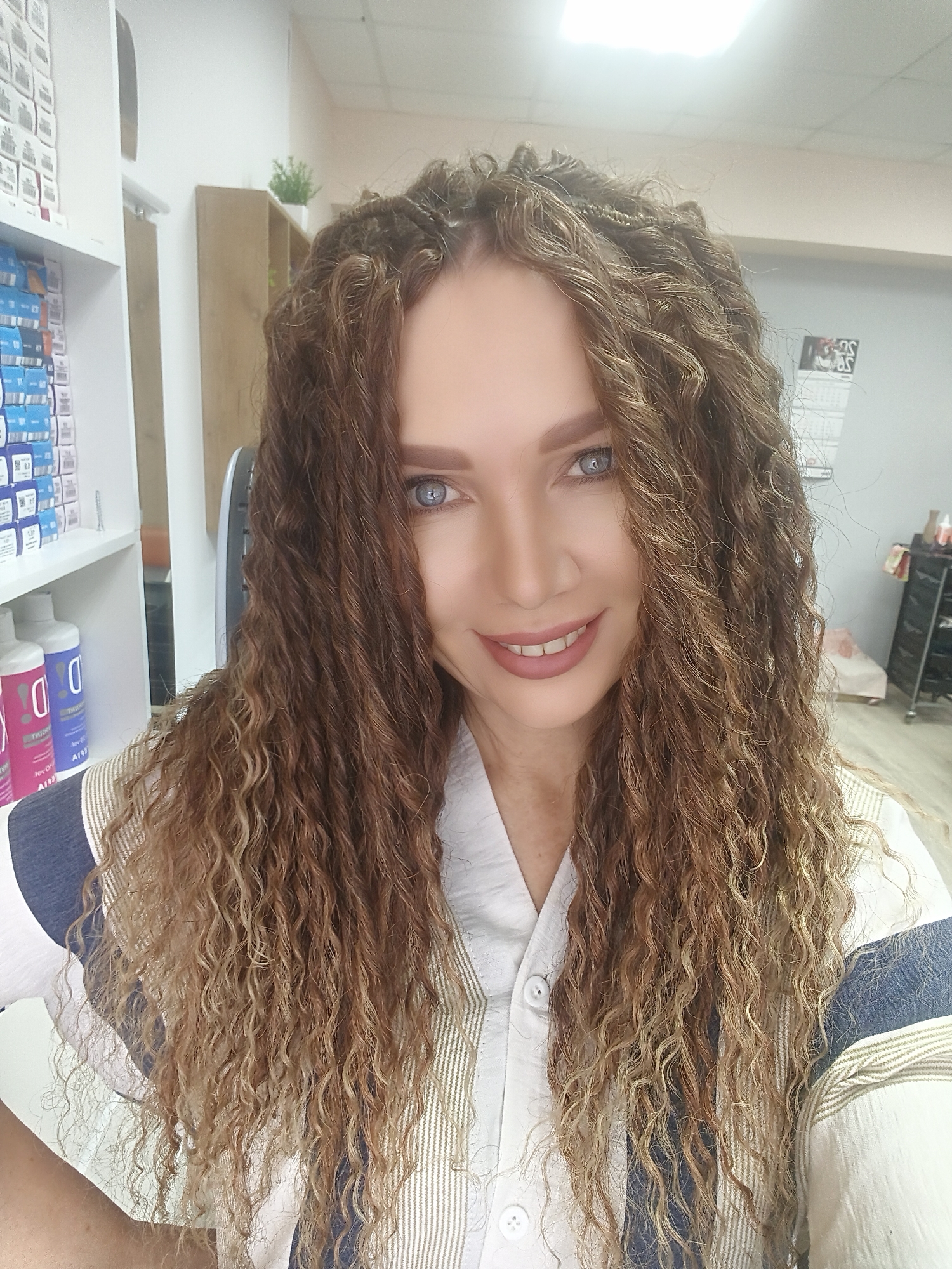 A woman with long, wavy to curly brown and blonde hair, and bright blue eyes, smiles directly at the camera in what appears to be an indoor commercial setting. She wears a white shirt with blue and light-brown striped sections. Her hair features some braided elements near the scalp. She is wearing light makeup, including pink lipstick.
The background on the left side of the image features white shelving units fully stocked with numerous small boxes, likely product packaging, and two larger bottles on a lower shelf, one pink and one blue, with visible text "DIO" and "AIYI" (partially obscured). On the right side, there is a light-colored wall, a wooden shelf holding a small green potted plant, and a calendar hanging on the wall with what appears to be "AS" visible. Further back and to the right, a dark cart or trolley with various items, and a light-colored piece of furniture are visible. Overhead, bright fluorescent lighting is present, indicating an interior space, likely a hair salon, beauty studio, or a retail store selling beauty products. The overall atmosphere conveyed by the woman's smile is friendly and inviting. No specific time of day is discernible, but the bright artificial lighting suggests an active business environment.