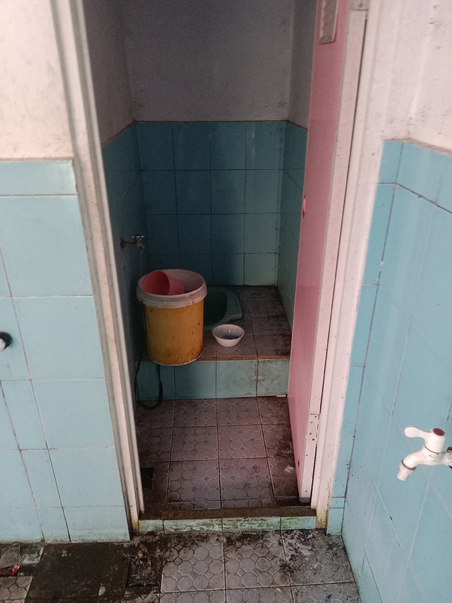 This image depicts a cramped, dimly lit bathroom stall with light blue tiled walls. Inside the stall, a yellow bucket with a red lid sits on a tiled platform. Next to the bucket, a small white bowl is visible. The floor of the stall is tiled with a mosaic pattern. A partially open pink door reveals the stall's interior. The area outside the stall appears to be a hallway or corridor with light blue tiled walls, a white faucet, and a grimy, tiled floor with visible debris. The overall condition of the bathroom suggests it is in a state of disrepair.