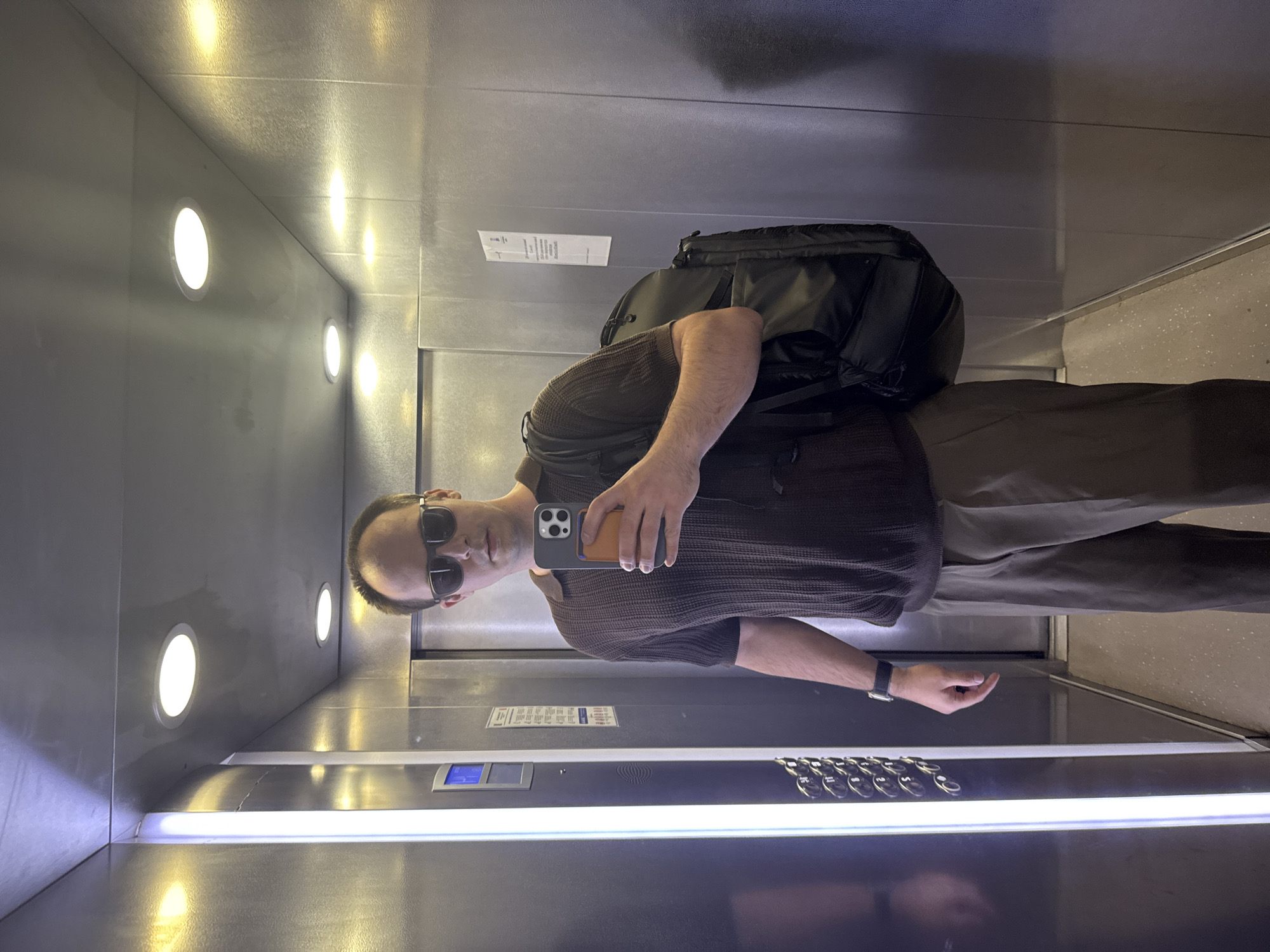 A man stands inside an elevator, facing the reflective wall where his image is visible. He is wearing sunglasses, a brown shirt, and brown pants, with a black backpack slung over his shoulder. He holds a cell phone up, seemingly taking a picture of himself. The elevator is illuminated by a series of circular lights recessed into the ceiling. An elevator control panel is seen. The environment is an elevator, but there is no forensic evidence to suggest this is in Perm, Russia.