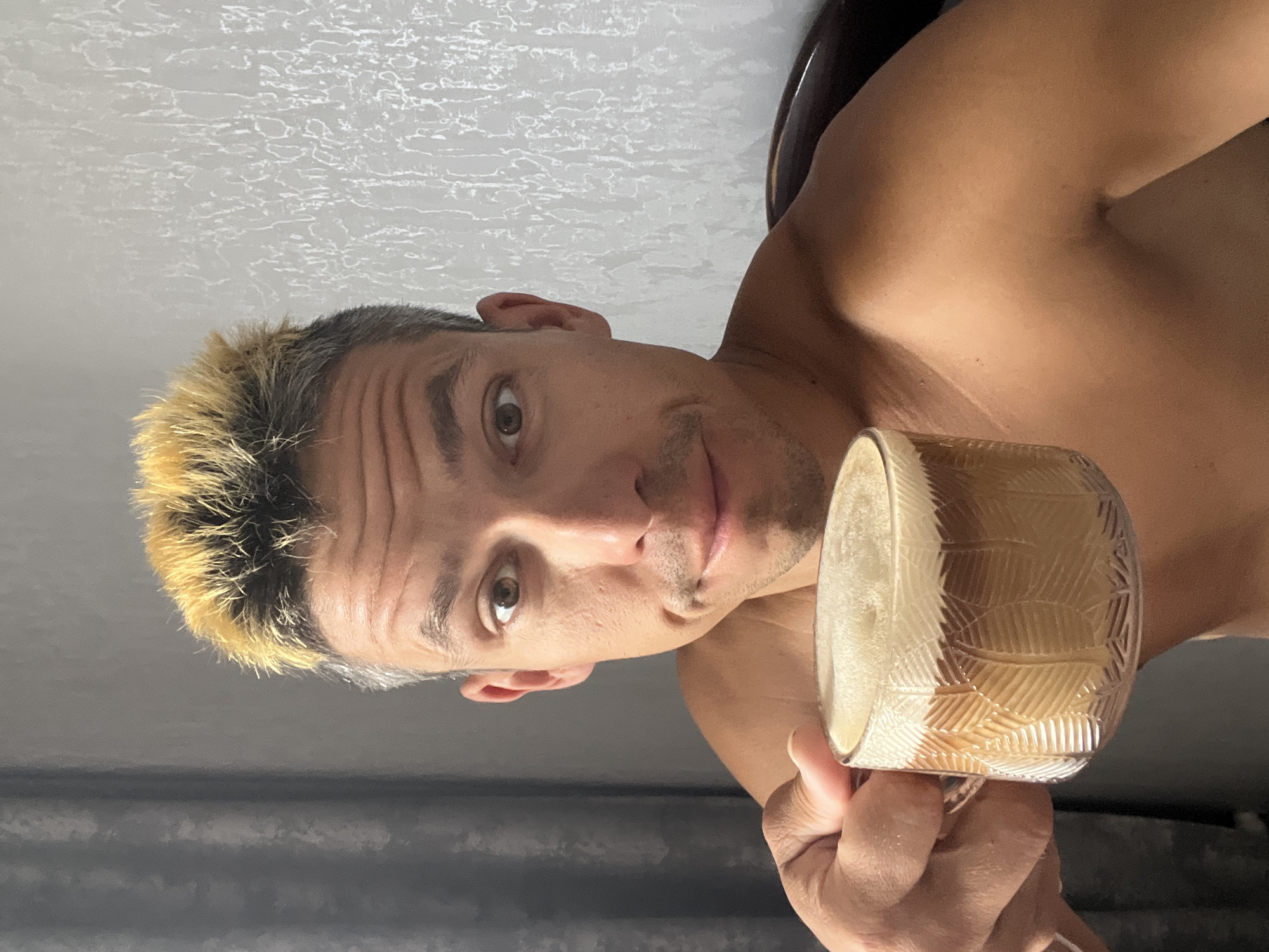 A man with short, spiked, bleached blonde hair and grey roots is holding a clear glass mug filled with a foamy, brown beverage. He is shirtless and looking directly at the camera with a slightly furrowed brow and a mild smile. The mug has a textured pattern of what appear to be leaves etched into the glass. The background consists of a grey, textured wall and a dark curtain. The scene is dimly lit, suggesting an indoor setting. The image does not contain any visual information that would allow for the identification of its specific location within the city of Chaikovsky, Russia.