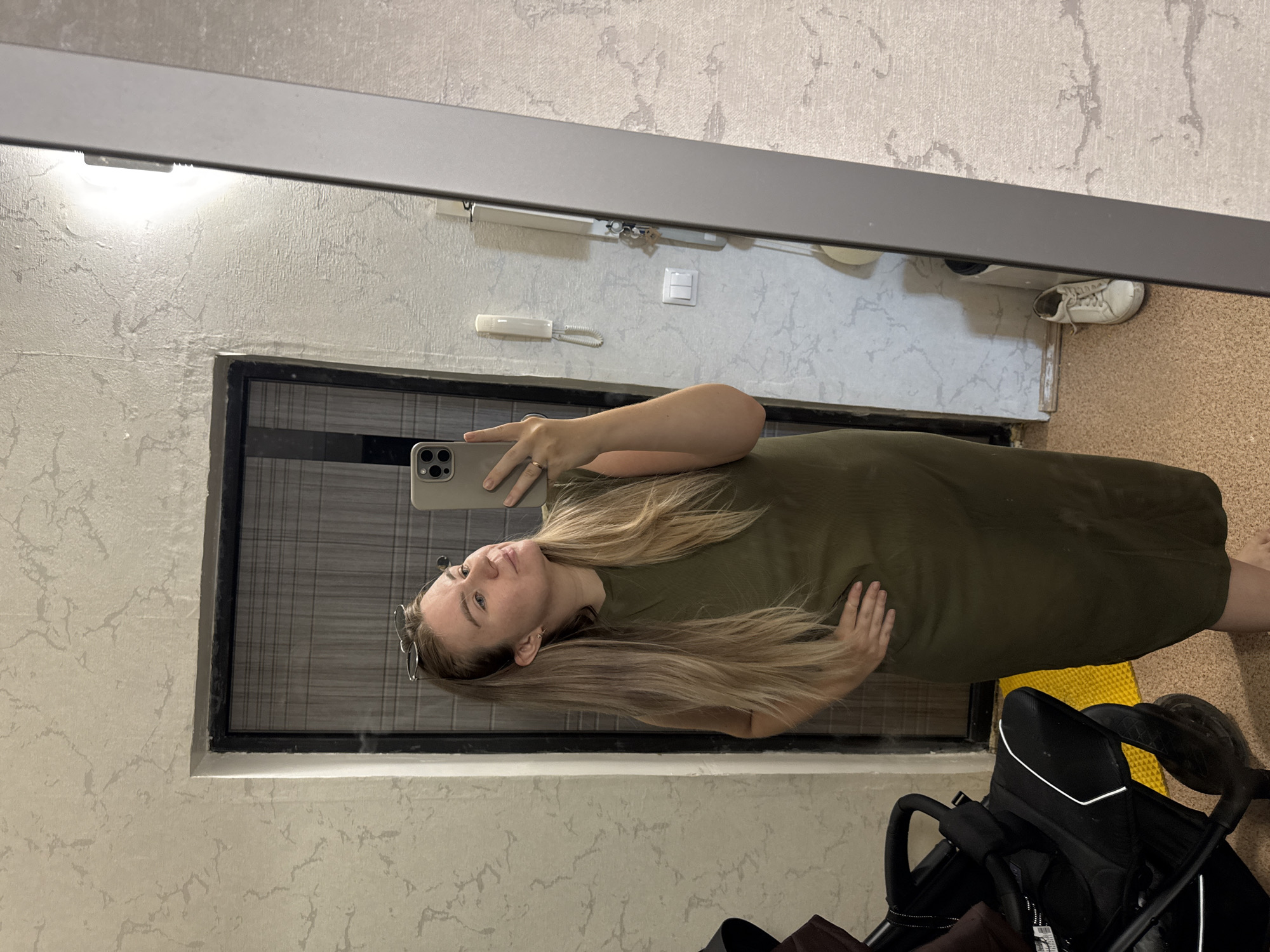 The image shows a woman taking a selfie in a mirror. She is wearing a green dress and has long blonde hair. A stroller is partially visible in the bottom right corner of the image. The background includes a door, an intercom system, and a shoe. The location is Chaikovsky, Russia, based on the information provided. The setting appears to be an apartment hallway.