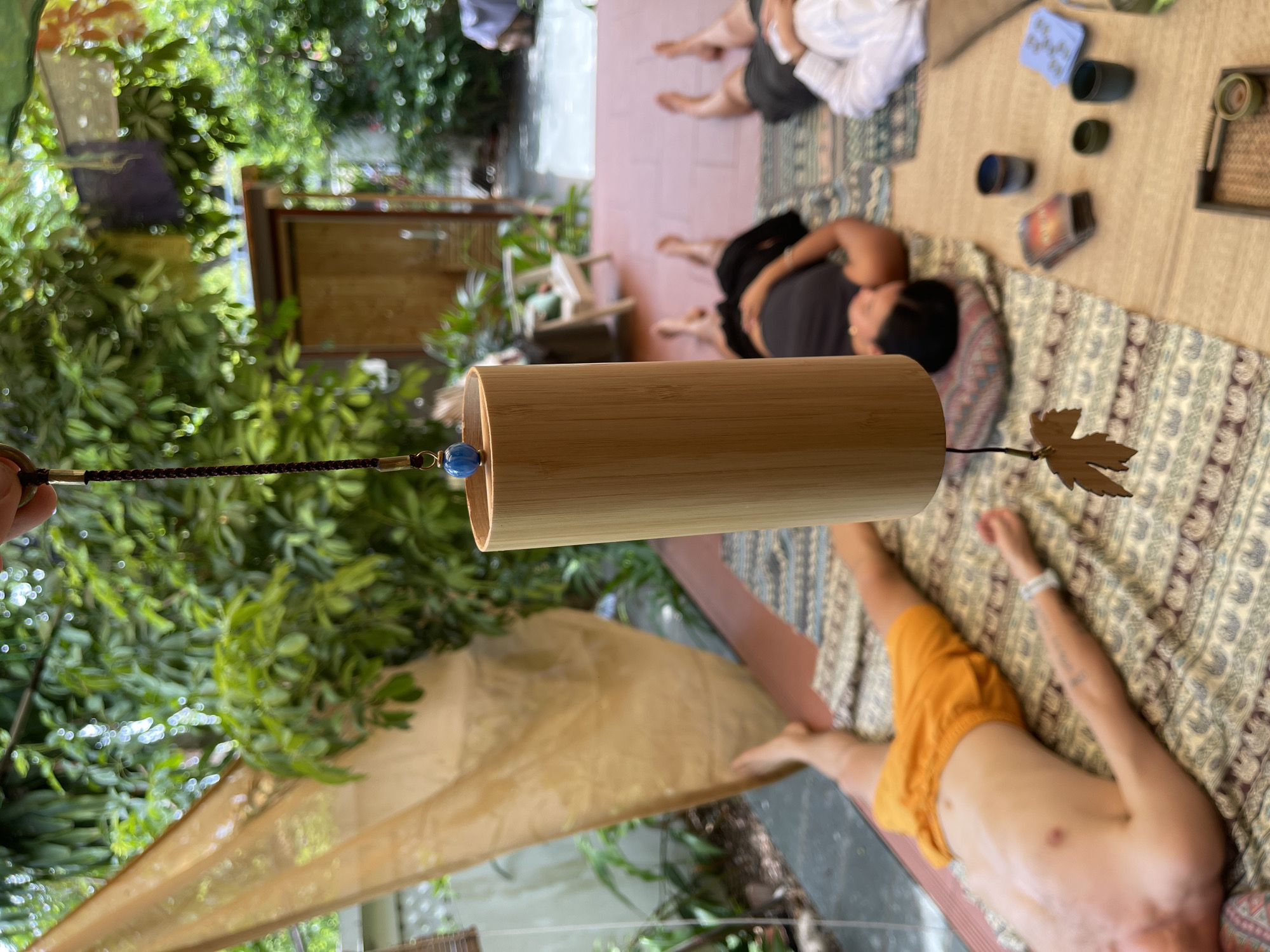 In Đà Nẵng, Vietnam, four individuals are seen lying on mats in what appears to be an outdoor or semi-outdoor space. The setting is lush with abundant greenery, suggesting a garden or courtyard. In the foreground, a bamboo wind chime hangs suspended by a braided cord, adorned with a blue bead and a wooden leaf charm. The wind chime is positioned centrally and is in focus.
To the left, a person lies on their back, wearing orange shorts. Their body is partially illuminated by sunlight. Further back, two individuals are lying on their backs on patterned mats. One is wearing a dark top, and the other is wearing a white top. Their heads are resting on pillows, and their legs are extended. A fourth person, partially visible, is also lying down.
Scattered on a mat near the center right are several small cups, a stack of cards, and a small green object within a dark frame. The ground is covered with tiles and mats, and a beige fabric is draped on a structure to the left. The background reveals more vegetation and architectural elements, hinting at a residential or retreat-like environment. The overall atmosphere suggests a moment of relaxation or meditation in a tropical setting.