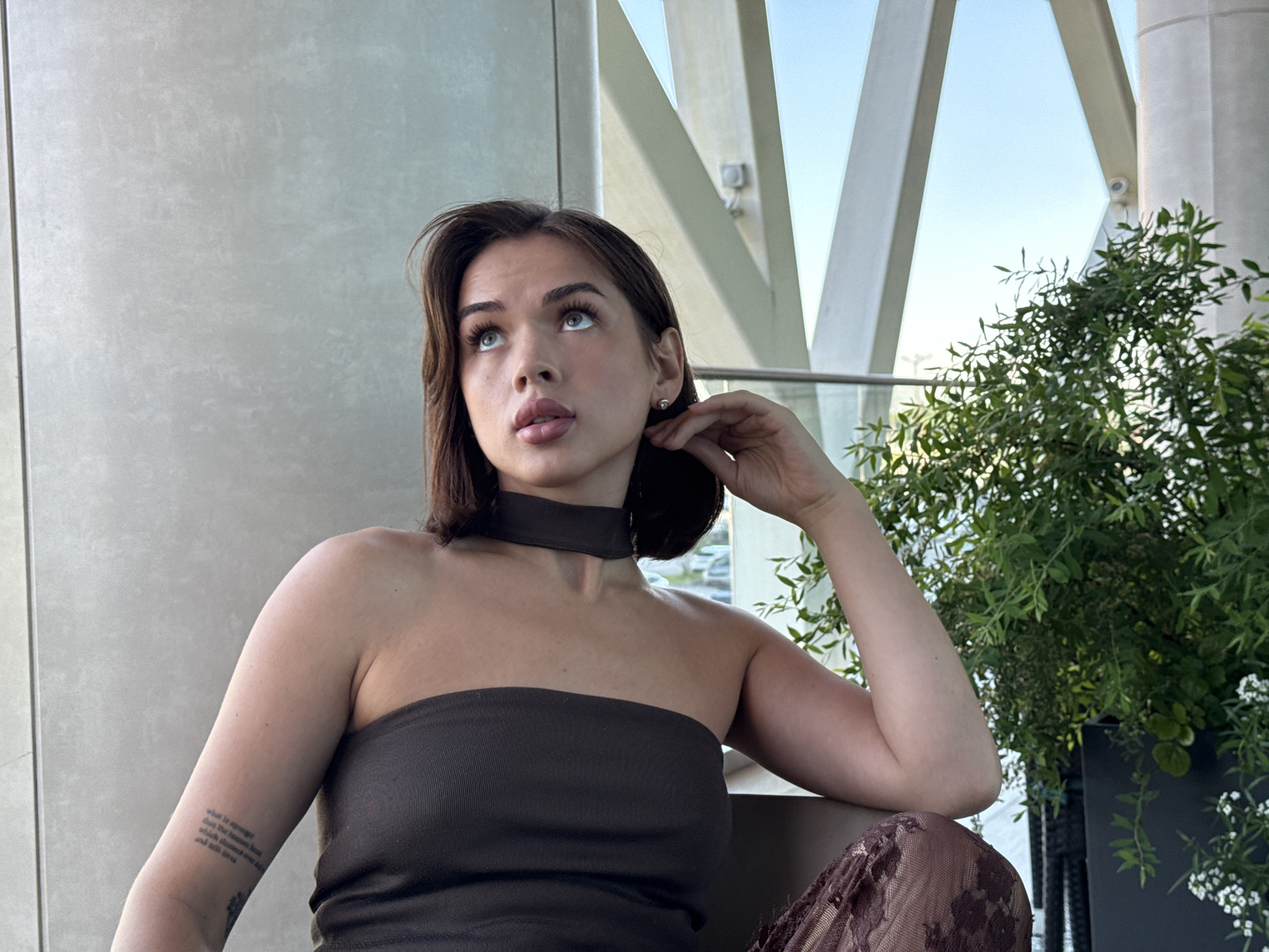 A young woman with medium-length brown hair is looking upwards and to her left. She is wearing a strapless dark brown top and a dark choker necklace. Her left arm is resting on her leg, and her right hand is touching her ear. A tattoo is visible on her left arm. The background shows a grey wall, a window with a view of the sky and white architectural beams, and a potted green plant. The image appears to be taken indoors with natural light. There is no indication of the location being the city of Perm, Russia.