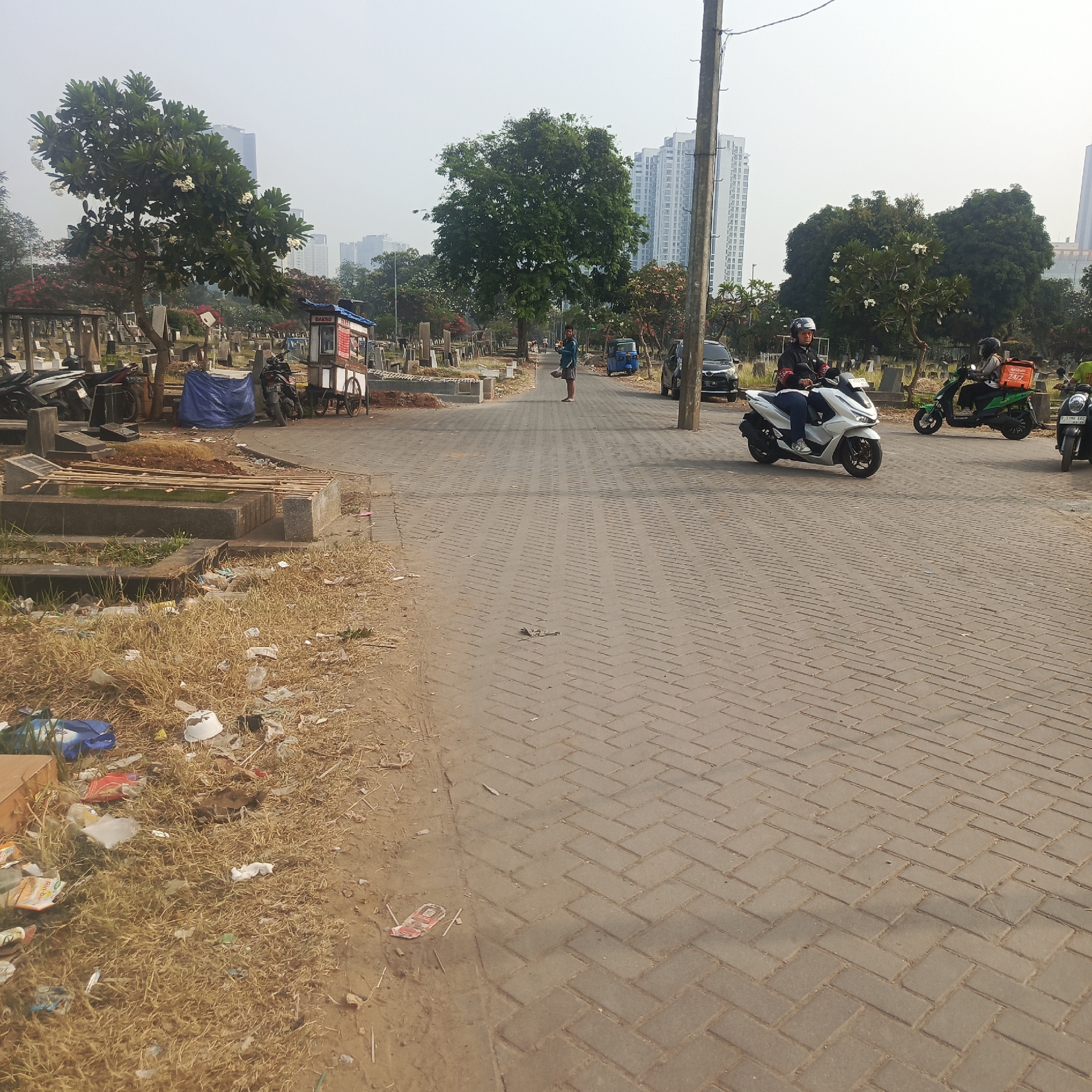 The image depicts a cemetery in South Jakarta, Indonesia. In the foreground, the ground is covered in dry grass and scattered litter. A paved pathway, made of interlocking bricks laid in a herringbone pattern, runs through the cemetery. Several gravestones and burial plots are visible on either side of the path. In the middle ground, a person is walking away from the camera, carrying a basket. Several motorcycles are parked along the path. Further down the path, two people are riding motorcycles. One is on a white scooter, and the other is on a green scooter with an orange box on the back. Tall apartment buildings can be seen in the background. The sky is overcast.