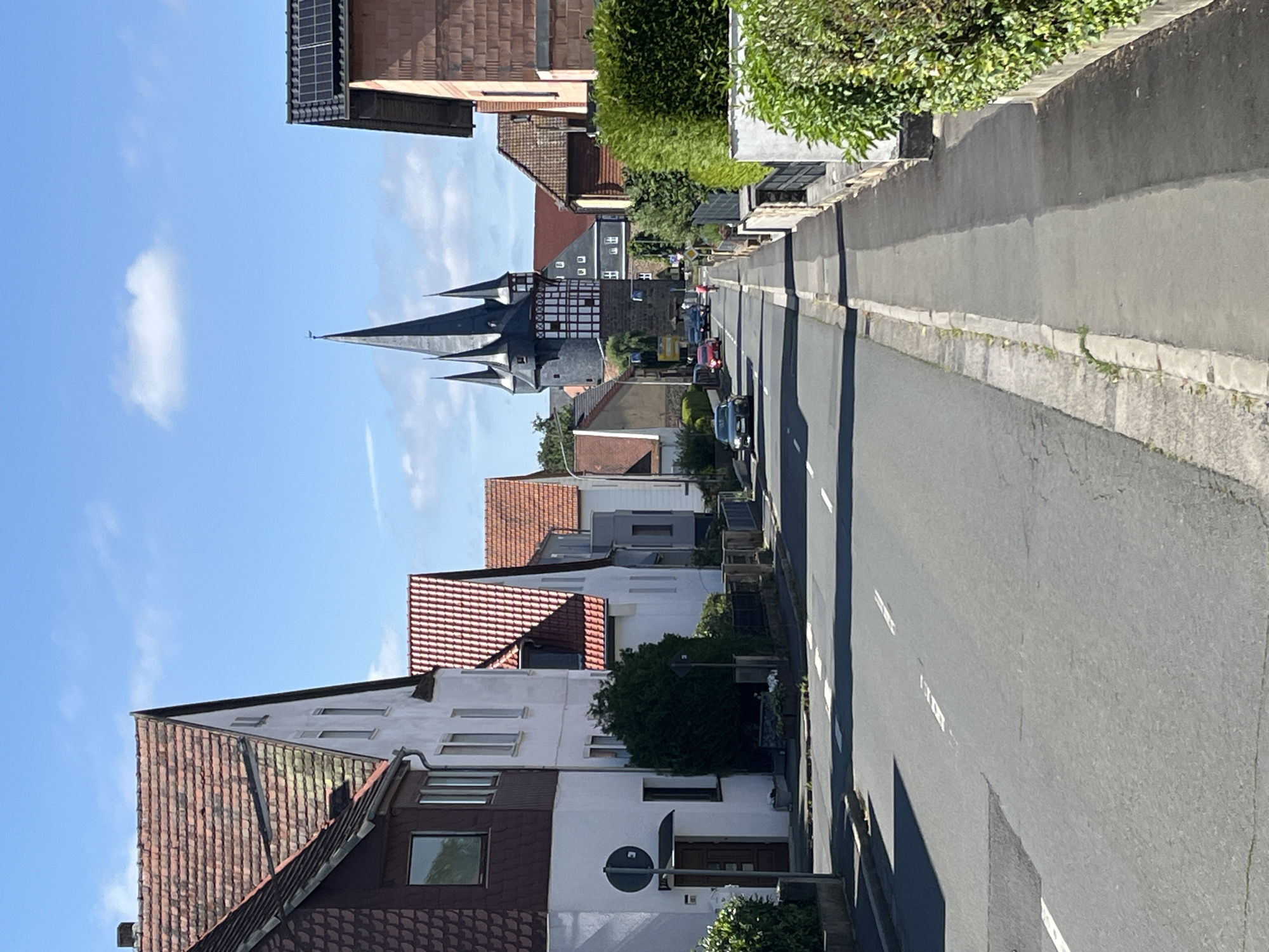 This is a daytime, eye-level, full shot of a street scene in Neustadt, Germany. The road is asphalt with white lane markings. On the left side of the street, there are several houses with red tile roofs. One house is white with dark brown trim around the windows and a dark brown mansard roof. Another house is white with a red tile roof and a darker section above its window. A large green bush is in front of the white house. On the right side of the street, there is a sidewalk with a low stone wall and a hedge behind it. Further down the street, a large tower with multiple spires and a half-timbered section is visible. Several cars are driving on the road, including a black car on the left and red cars in the distance. The sky is blue with a few white clouds.