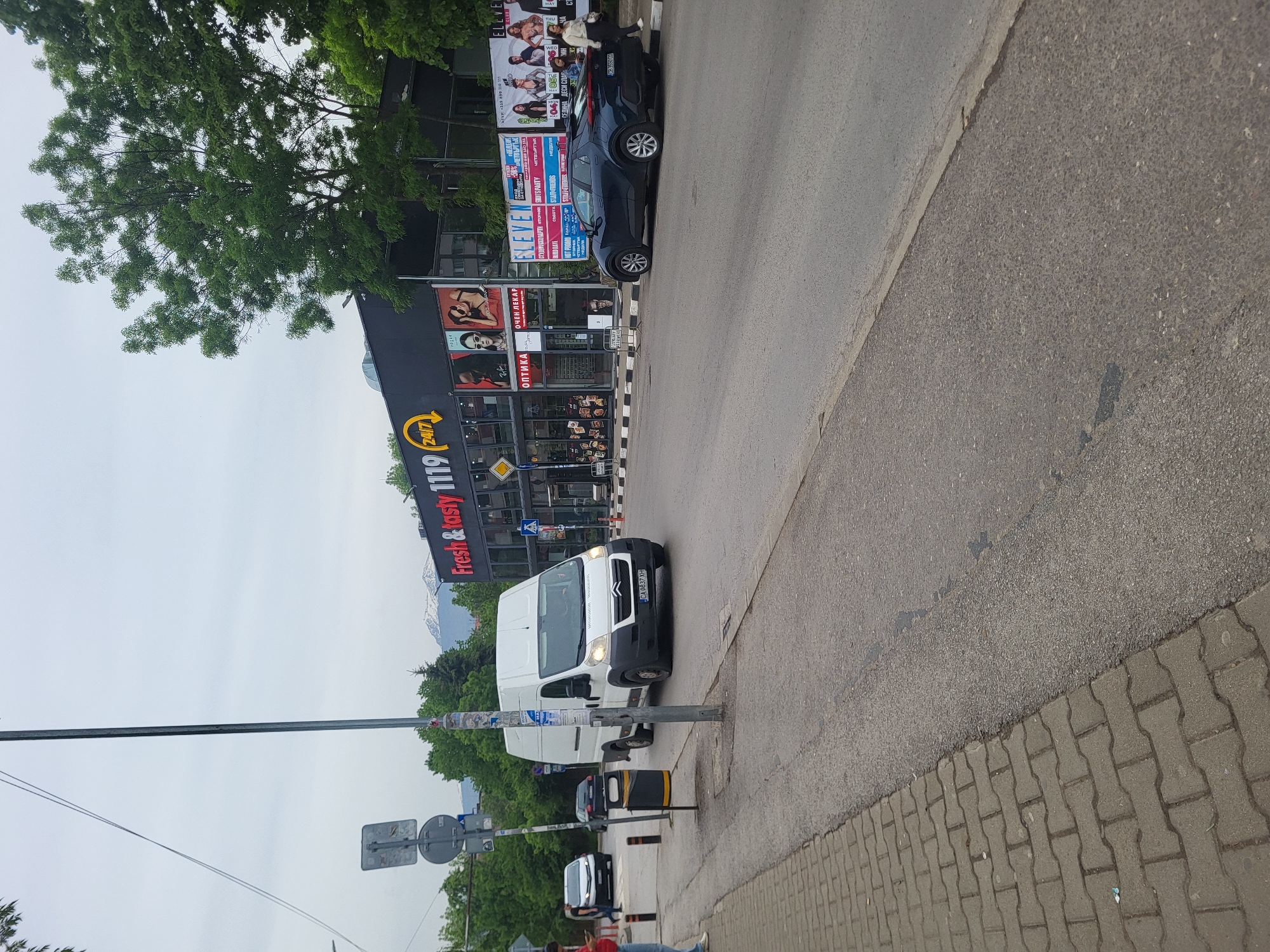 The image shows a street scene in Sofia, Bulgaria. A white van with license plate "CA 0837 XH" is parked near the sidewalk. Behind the van, a traffic signal post and other vehicles are visible. The background includes a building with a "Fresh & tasty 1119 24/7" sign. Several advertisements are visible on the building, including one for an optician ("ОПТИКА ОЧЕН ЛЕКАР") and another advertising "ELEVE," a club with event details. Mountains can be seen in the distance. The street is paved, and the sidewalk is made of interlocking bricks. There are trees and greenery along the street, and the sky is overcast.
