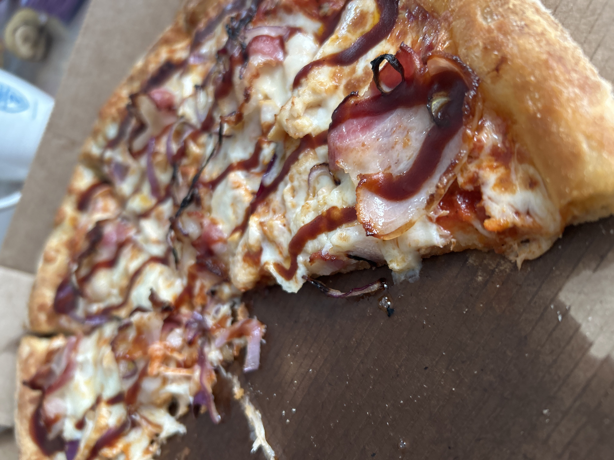 The image shows a partially eaten pizza in a cardboard box. The pizza appears to have a thick crust and is topped with cheese, bacon, red onions, and a dark brown sauce, possibly barbecue sauce. The pizza is sliced, and one piece is partially removed. The box is plain brown cardboard. The background is slightly out of focus. It is not possible to determine the location of the image as Chaikovsky, Russia from visual evidence.