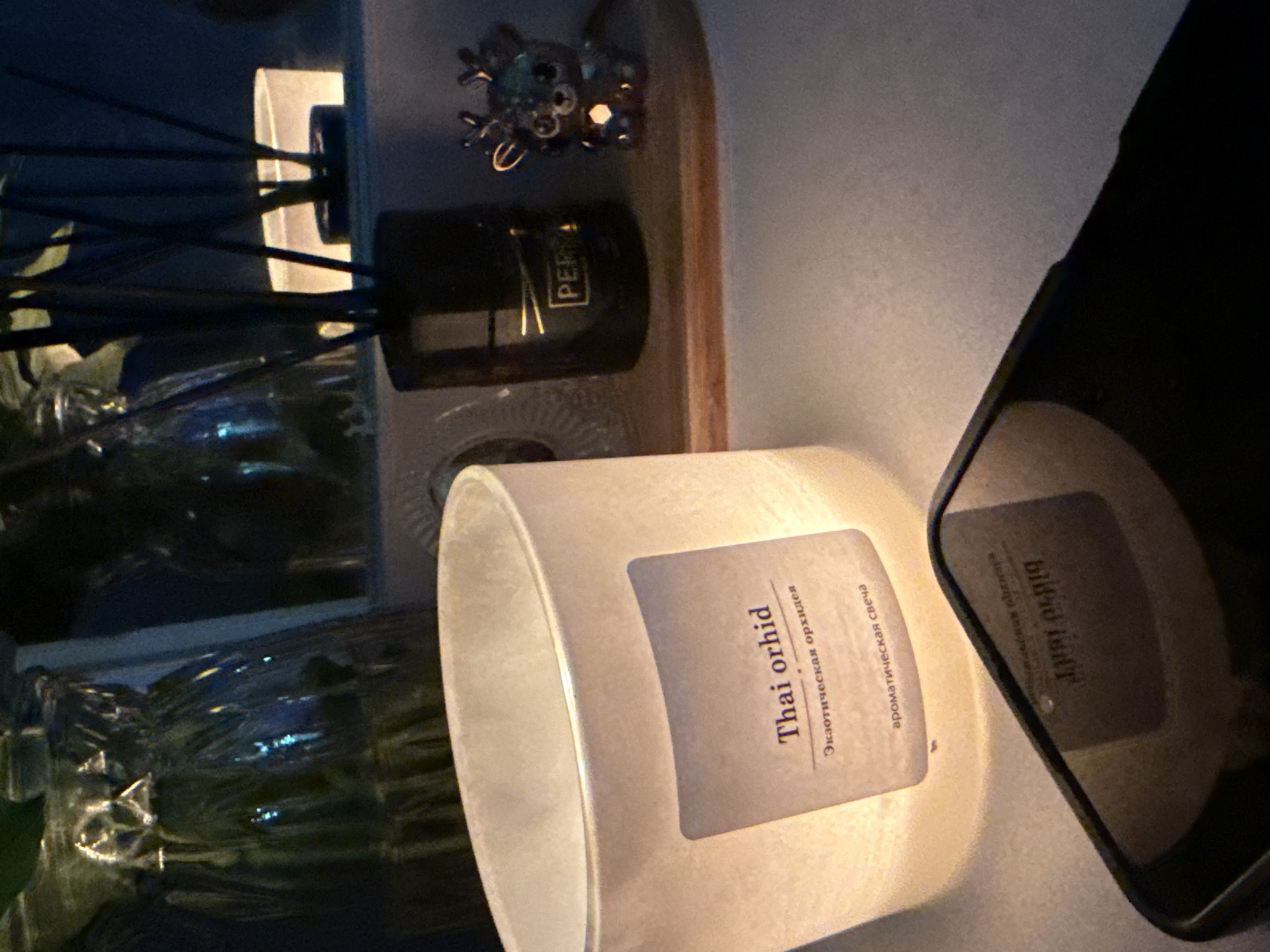 A close-up shot captures a dimly lit indoor scene with a focus on a scented candle. The candle, made of white frosted glass, emits a warm glow from within, illuminating the label. The label displays the text "Thai orhid" in English, followed by "Экзотическая орхидея" in Russian, and "ароматическая свеча" also in Russian. To the right of the candle, a black diffuser with reeds is visible, and further to the right is a small, decorative reindeer figurine. In the foreground, a portion of a dark smartphone is visible, reflecting the ambient light and some of the surrounding objects. The background features the blurred shapes of glass vases or bottles, suggesting they might contain plants or decorative elements. The overall ambiance is cozy and relaxed. The image does not provide any specific visual cues to locate it in Oktyabrskoye, Russia.
