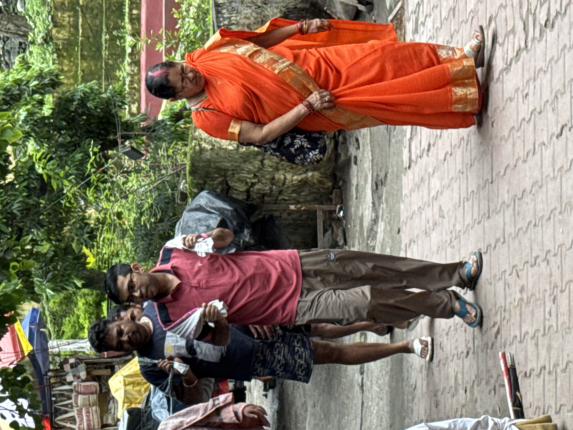 In Rishikesh, India, on a cloudy day, three people are walking along a patterned brick pathway. To the right, a woman in an orange sari with gold trim walks towards the foreground, carrying a dark patterned bag over her left shoulder. She wears glasses and jewelry, including bangles on both wrists. In the middle ground, a man wearing glasses, a pink polo shirt, and brown pants walks forward, holding a white cloth in each hand. To his left, another man in a dark t-shirt and patterned shorts walks forward, also holding a white cloth. Behind them, a person in a pinkish jacket is partially visible. The background features lush green trees, a wall, and stalls with various items, suggesting a market or street scene.