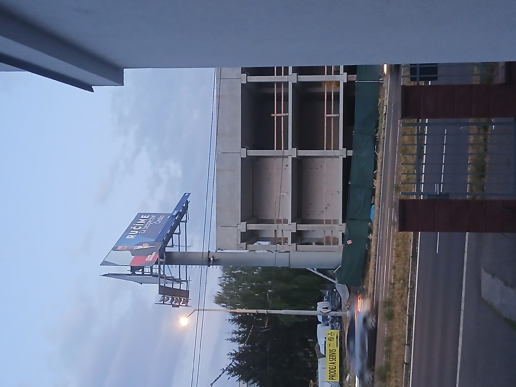 The image shows a construction site with a partially built concrete structure. In the foreground, a street with a car is visible. In the background, a billboard with text in Czech and an image of a person is displayed on a tall pole. The sky is overcast, suggesting it might be early morning or late evening. The construction site appears to be located in Pilsen, Czechia, as indicated by the billboard which reads "RÚČÍME za bezpečné Česko" (We guarantee a safe Czechia), a slogan often associated with political campaigns in the Czech Republic.