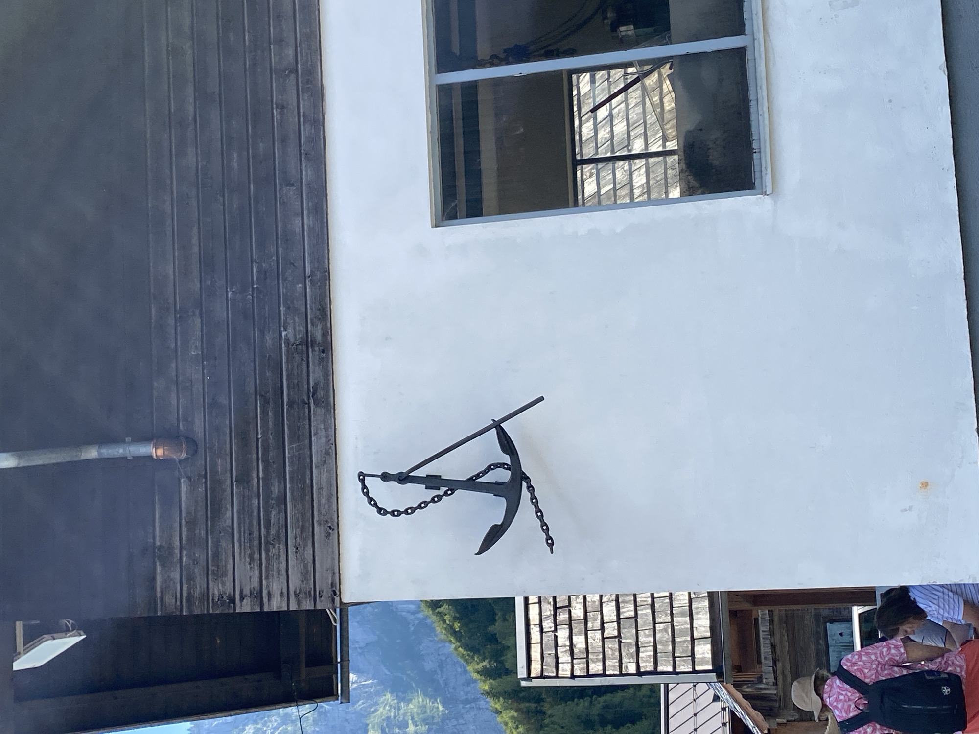 A black anchor with a chain is mounted on the side of a white building. The anchor is decorative and appears to be made of metal. Behind the white building, a wooden-clad structure with a shingled roof is visible, and further in the background, a mountainside covered in trees can be seen. To the right of the anchor, a large window with reflections is present. In the lower left corner, a person wearing a pink and white patterned shirt and a straw hat is visible, facing away from the camera, with a black backpack. Another person is partially visible next to them. The scene appears to be in an outdoor, possibly tourist, setting in Hallstatt, Austria, given the architecture and natural landscape.
