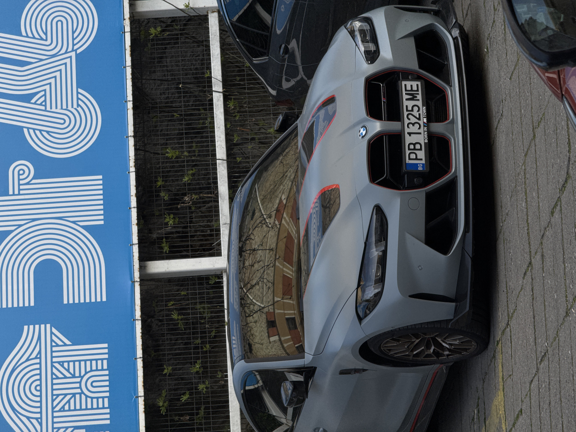 A gray BMW car, bearing the Bulgarian license plate "PB 1325 ME" and the inscription "BORN IN MTOWN," is parked on a brick-paved surface. A blue banner featuring a white geometric design hangs to the side. The car's windshield reflects the image of a building. The presence of Bulgarian symbols on the license plate suggests that the image was taken in Bulgaria, but there is no definitive evidence it was taken in the city of Plovdiv.