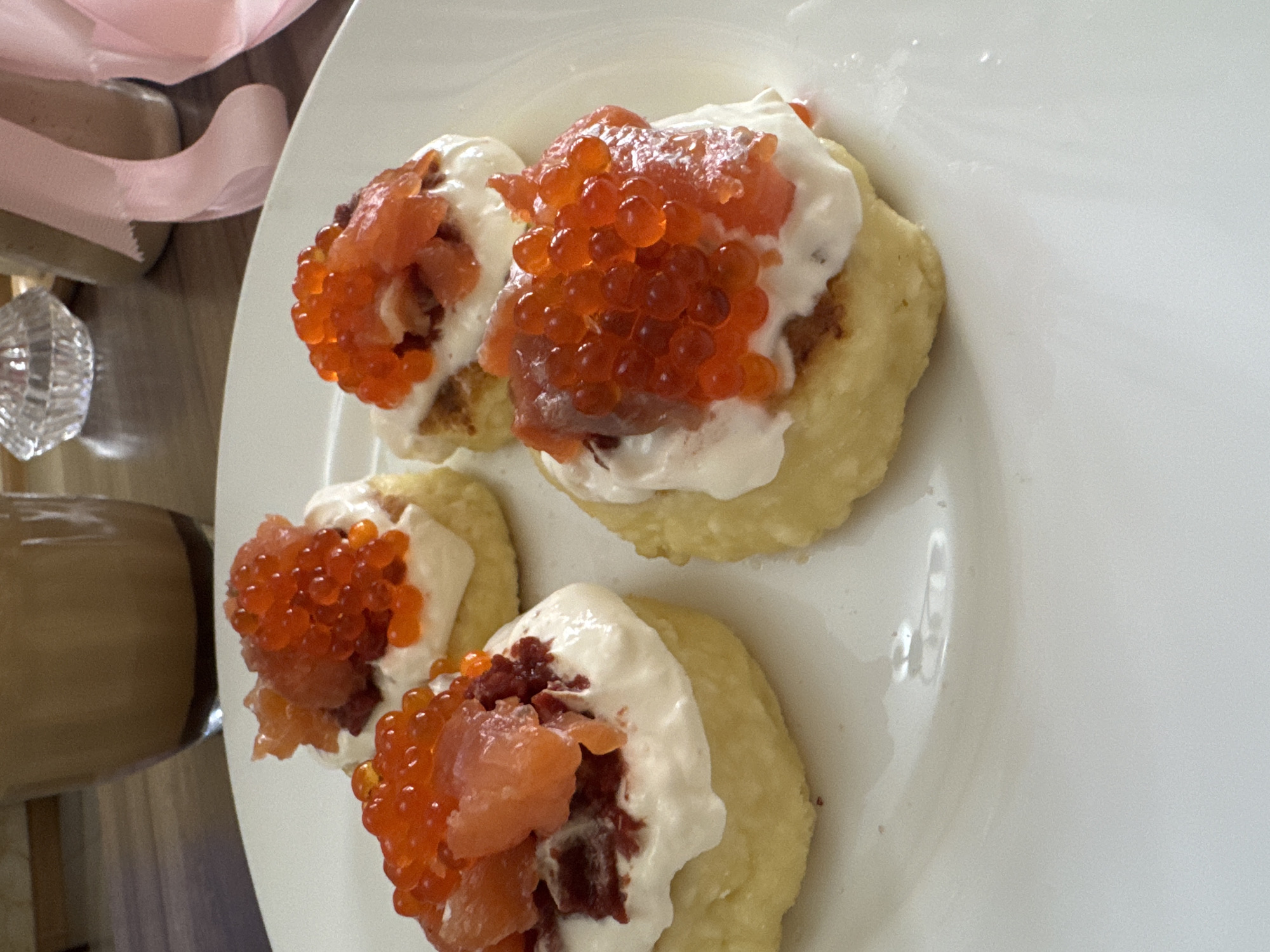 The image depicts a close-up shot of a white plate containing four small, round appetizers. Each appetizer consists of a base, possibly a blini or small pancake, topped with a white creamy layer and a generous portion of orange-colored fish roe or caviar. Some pieces of salmon also appear to be present on top of the creamy layer, mixed in with the roe. The plate is situated on a surface that includes a glass bottle and a light pink ribbon. The location and city are Unknown, Unknown.