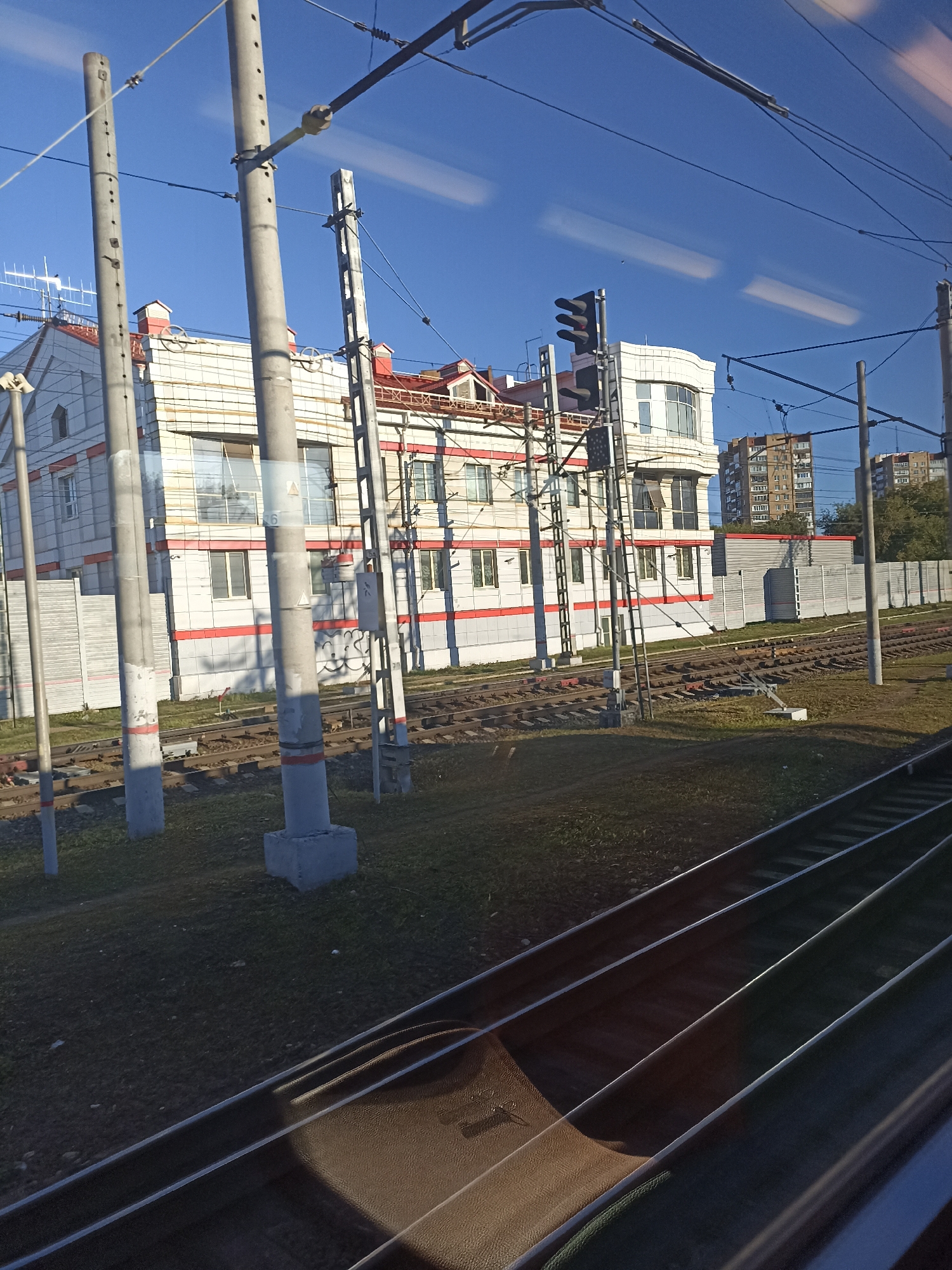 The image shows a view from a train window of a building and railway tracks on a clear day. The building is modern with white and red horizontal accents and several windows. It appears to be an industrial or commercial building, possibly a business center or a factory. There are railway tracks in the foreground and middle ground, with overhead power lines visible against the blue sky. In the background, more buildings and trees can be seen. The scene is likely located in an urban or suburban area, possibly on the outskirts of Podolsk, Russia, given the presence of railway infrastructure and multi-story buildings. There are no people visible in the image. The lighting suggests it is daytime.