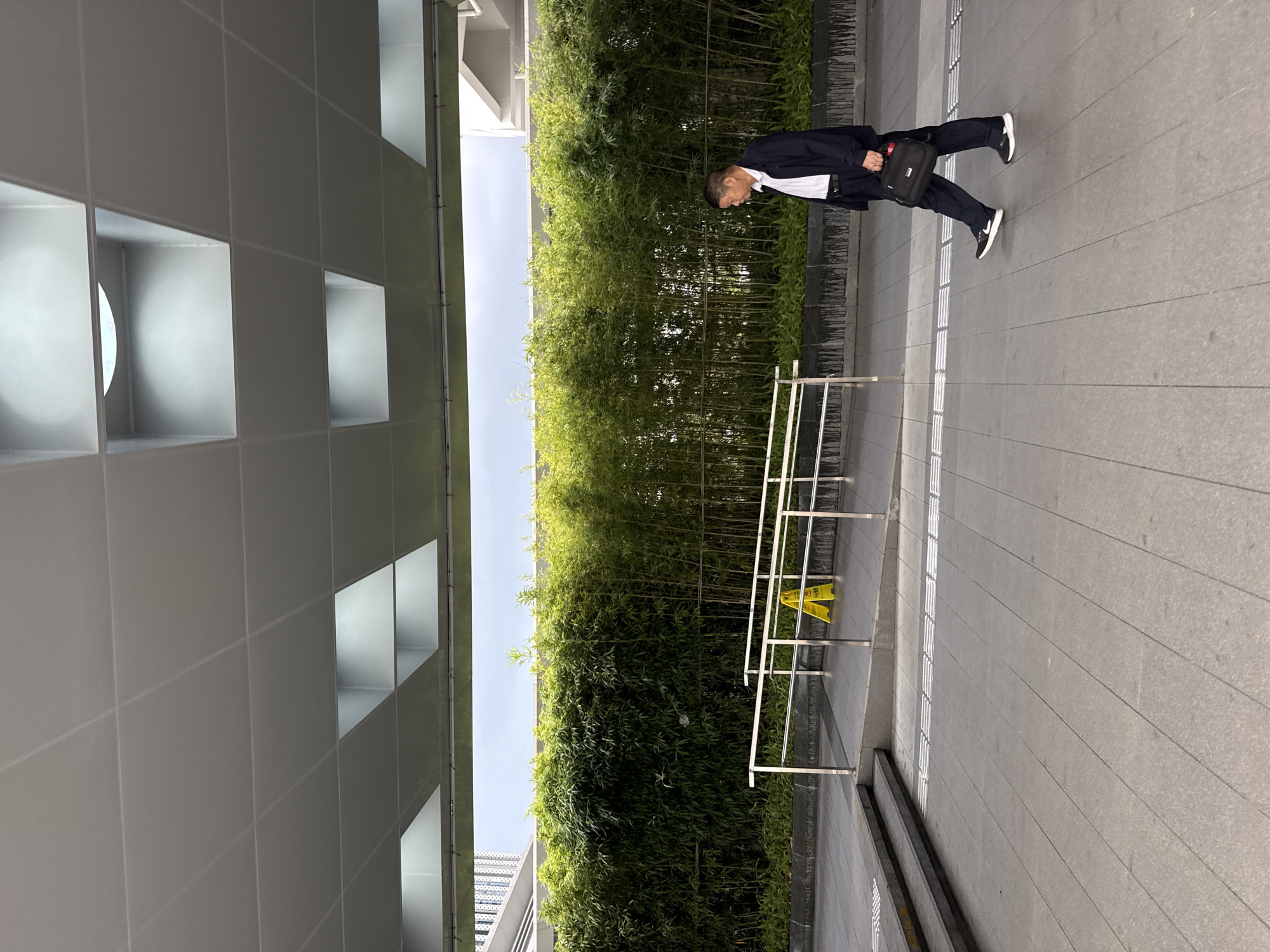 An East Asian man in a dark suit jacket, white shirt, and dark trousers walks on a paved outdoor walkway. He carries a black briefcase in his right hand. The walkway is bordered by a metal railing. Beyond the railing is a ramp leading to stairs. A yellow caution sign is visible on the ramp. Behind the railing and ramp is a dense wall of green bamboo plants. Above the scene is a modern architectural overhang with a grid of ceiling panels and recessed lighting. The sky appears overcast. The location is a city street in Huizhou, China.