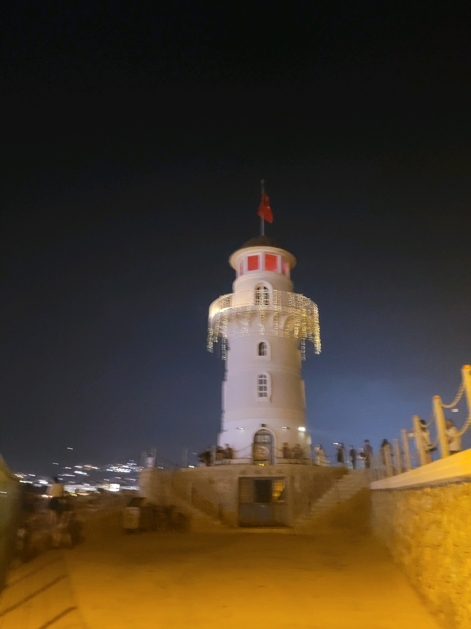 The image shows a lighthouse in Alanya, Turkey, at night. The lighthouse is white with red accents at the top and is adorned with festive string lights. A Turkish flag flies from the very top. The lighthouse stands on a stone foundation with steps leading up to its entrance. Several people are visible on the steps and near the lighthouse. In the background, the city lights of Alanya are visible against the dark night sky. The foreground shows a paved area with some indistinct figures and objects that appear to be vehicles. The overall scene suggests a lively atmosphere, possibly during a festival or celebration.