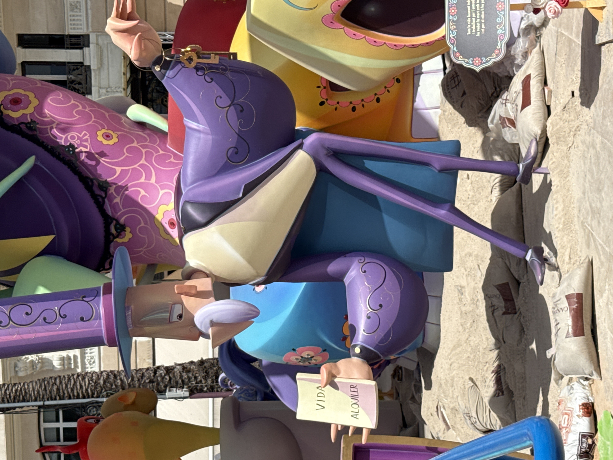 The image depicts a brightly colored, cartoonish sculpture likely from a street festival or public art installation. The sculpture features exaggerated, stylized figures and patterns. In the foreground, a character in a purple suit and hat is shown holding a small booklet. The booklet has text that reads "VIDA" and "ALQUILER." The character's left hand is extended, holding the booklet. This character is standing on a surface that appears to be sand, with several sandbags scattered around the base of the sculpture. Behind this main figure, other colorful, abstract sculptural elements are visible, including a large, curved shape in pink with floral patterns and a bright yellow element. The background shows a building with windows and balconies, suggesting an urban setting. The lighting indicates it is daytime.