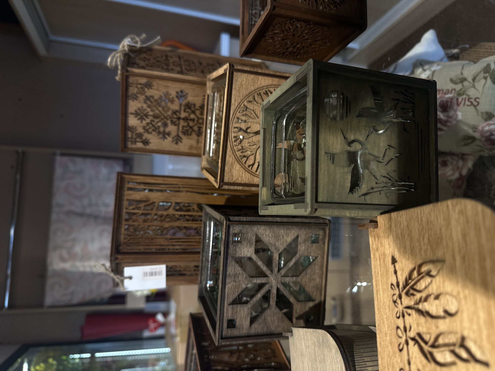 The image shows a close-up of stacked, decorative wooden boxes, likely for sale. The boxes feature various designs, including snowflakes, trees, birds, and feathers. Some have glass inserts. The scene is inside a shop, possibly a souvenir or gift shop. While the location is stated as Krustiņi, Latvia, there's no visual confirmation within the image.