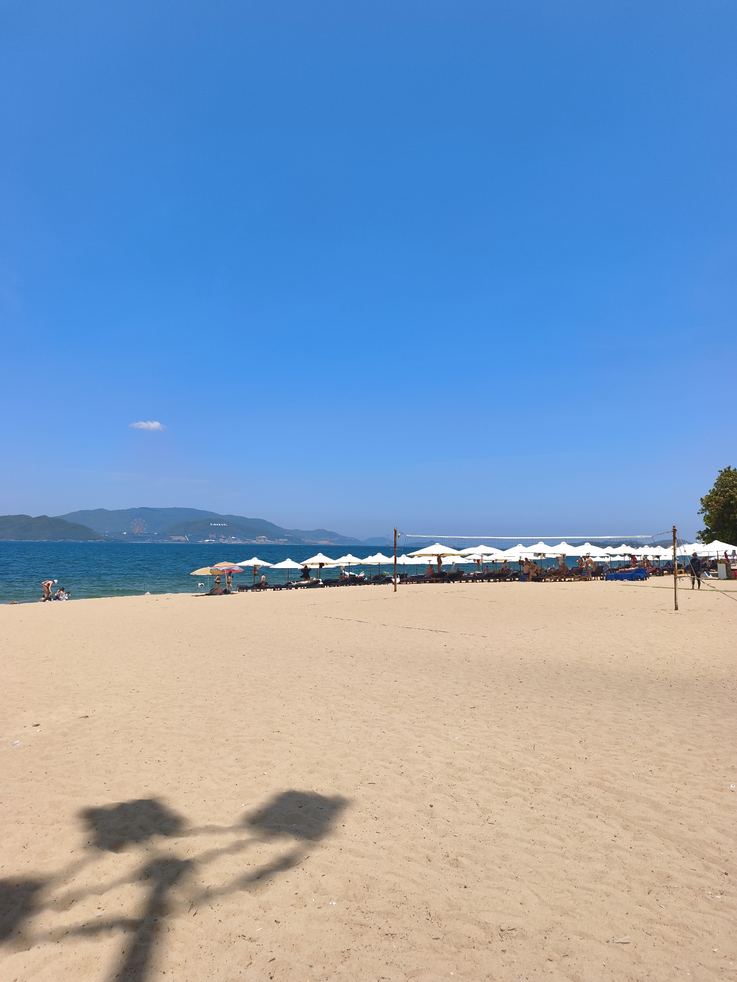 This is a wide-angle shot of a sandy beach in Nha Trang, Vietnam, under a clear blue sky. In the foreground, the sand is mostly empty, with two distinct shadows of what appear to be lamp posts or trees cast across it.
The middle ground features a line of white beach umbrellas and lounge chairs, some occupied by people. The water is a vibrant blue, meeting the sandy shore. In the distance, a chain of green, mountainous islands is visible, with a Ferris wheel and some buildings on the largest island. There are a few people scattered on the beach and in the water closer to the left side of the frame. A volleyball net is set up further down the beach, with people near it. The overall scene depicts a typical day at a tropical beach resort.