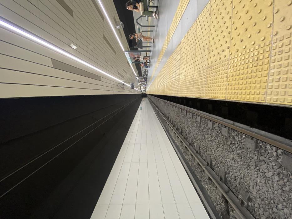 The image depicts an underground subway station, specifically a platform and track area, located within Line H of the Buenos Aires Subte, Argentina. The perspective is a low-angle, wide shot looking down the length of the station.
On the left, a single train track is visible, resting on a bed of gray ballast and wooden sleepers. The track runs parallel to a long, white wall constructed of horizontal, rectangular panels. Above the track and wall, the ceiling is uniformly dark gray or black.
On the right, a subway platform extends into the distance. The platform floor features distinctive bright yellow tactile paving tiles with both square and circular raised patterns. The non-tactile sections of the platform floor are light gray. Above the platform, the ceiling is a lighter color, likely white or light gray, with linear accents and integrated, continuous LED strip lighting running parallel to the platform's edge.
The dark ceiling area over the track visually contrasts sharply with the lighter, illuminated ceiling over the platform, meeting precisely along the vertical plane of the platform edge.
Several individuals are present on the platform. In the mid-ground, approximately three people stand near an indistinct information display or barrier. One individual in a white top and dark bottoms appears to be looking at a phone. Further along the platform to the right, a person with dark hair and sunglasses, wearing a light green shirt and khaki pants, is seated on a metallic bench. Other figures are visible further down the platform, some seated on benches, others standing, waiting. No train is present in the station. The overall environment appears clean and modern.