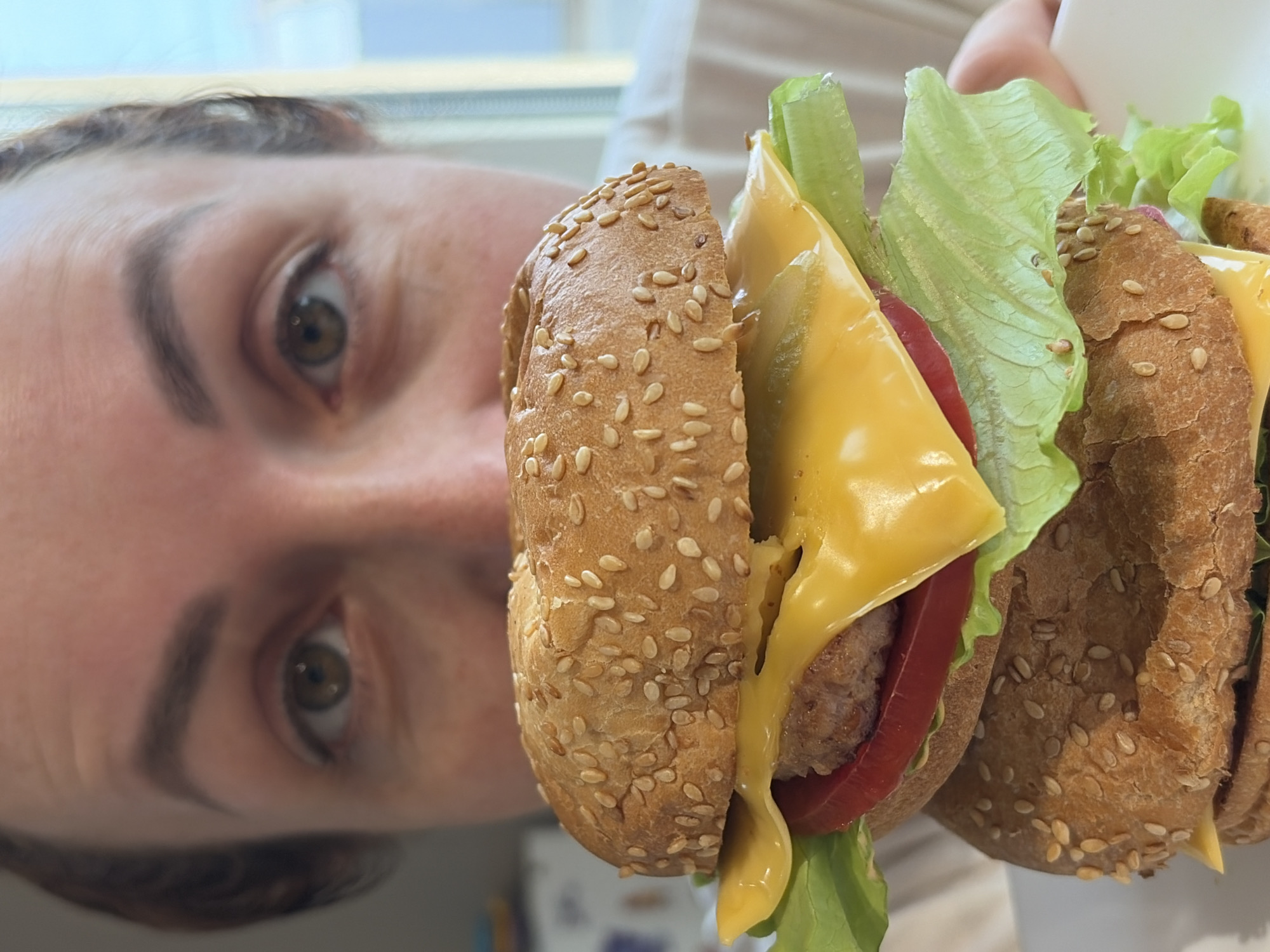 The image shows a person holding a burger in front of their face. The burger appears to have a sesame seed bun, cheese, a slice of tomato, and lettuce. The person's face is visible on the left side of the image with their eyes looking towards the camera. The location is identified as Chaikovsky, Russia.