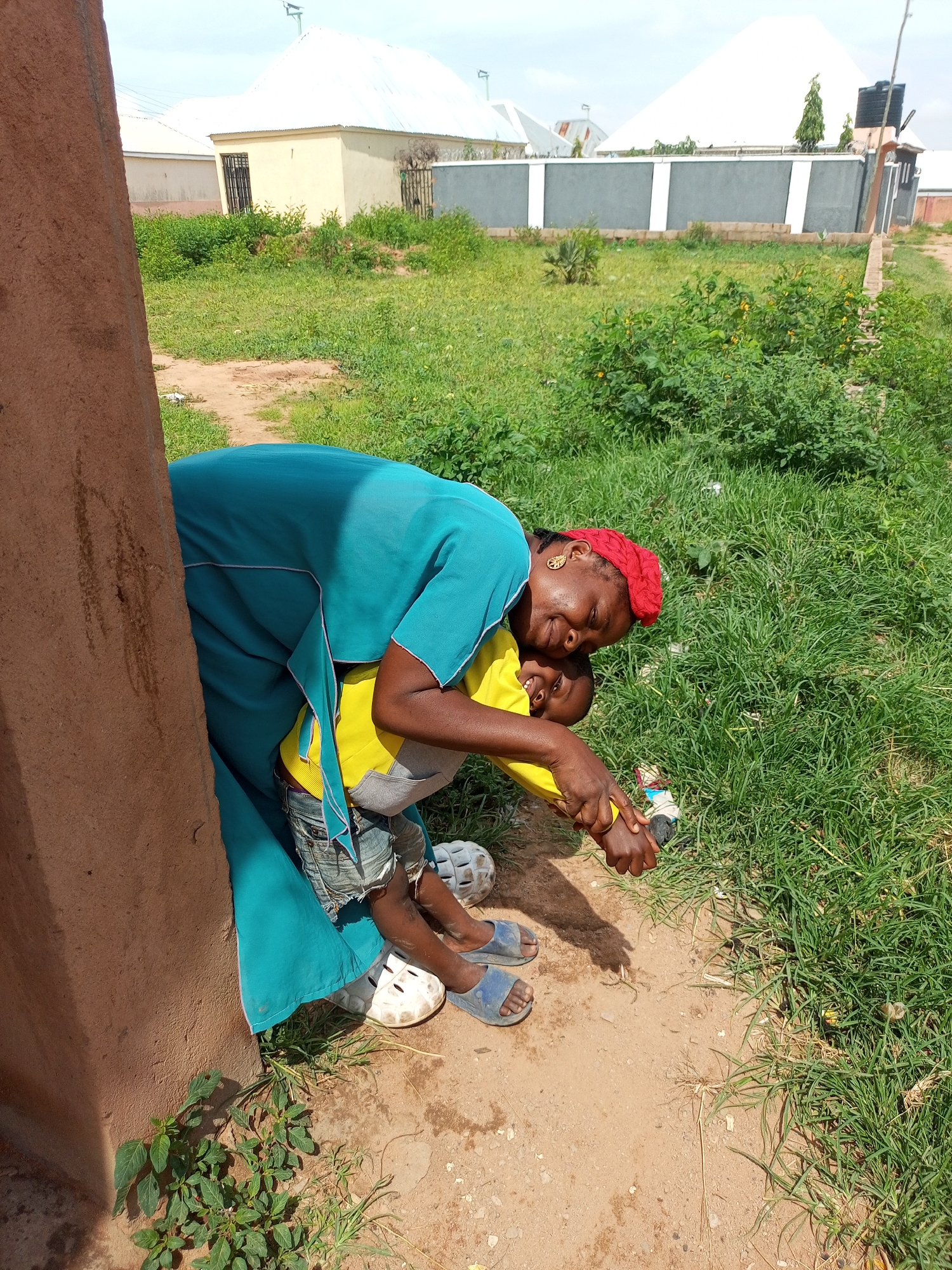 A woman wearing a teal-colored dress and a red headscarf embraces a young boy dressed in a yellow t-shirt and denim shorts. They are standing outdoors, leaning against a brown, textured wall. The ground is a mixture of dirt and sparse green grass. In the background, several buildings are visible, including houses with white roofs and grey-and-white patterned walls. The overall scene suggests a residential area in the city of Tempure, Nigeria.