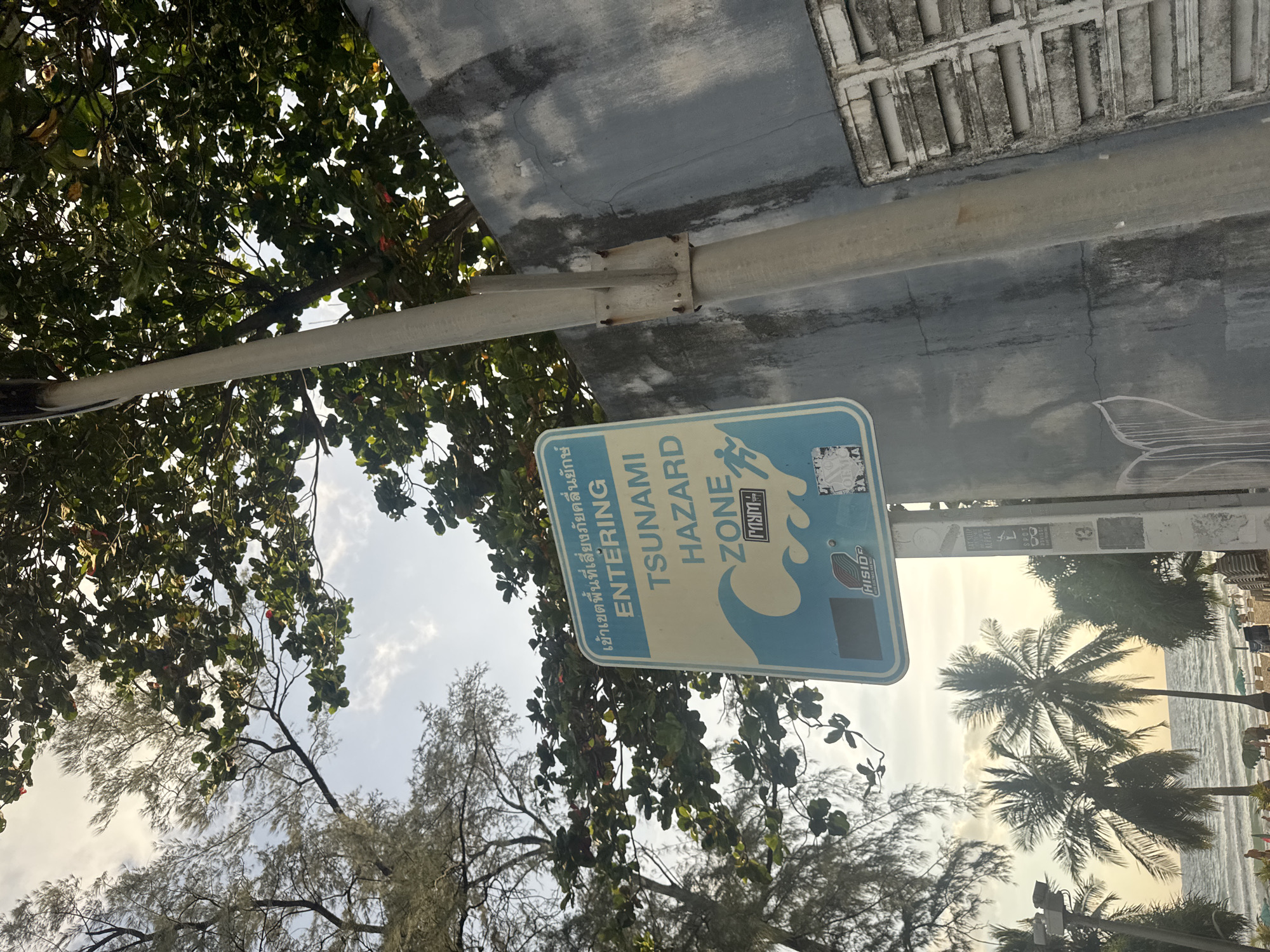 A tsunami hazard zone sign is mounted on a pole on the left side of the image. The sign is blue and white, with text in Thai and English. The English text reads "TSUNAMI HAZARD ZONE". The sign also features a wave graphic and a silhouette of a person running. The pole is gray and made of metal. Behind the sign, there is a gray concrete wall with some water stains and graffiti. Trees with green leaves are visible above and behind the sign. To the right of the pole, a section of a wall with horizontal vents is visible. In the background, palm trees and a beach with umbrellas and people can be seen, with the ocean and a hazy sky in the distance. The image appears to have been taken in Karon, Thailand.