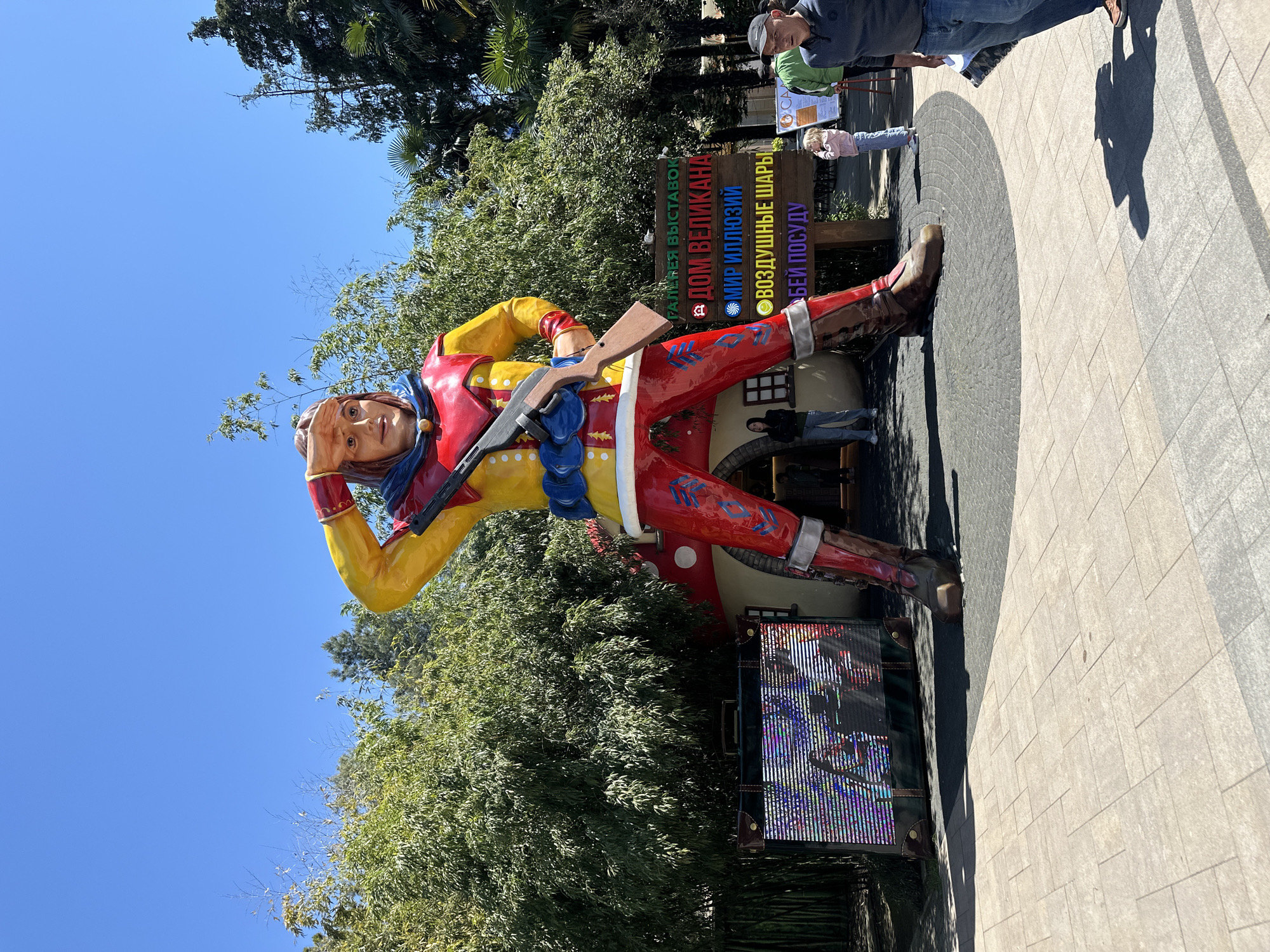The image displays a vibrant outdoor scene in Sochi, Russia, centered around a large, colorful statue of a male figure. The colossal statue, resembling a cowboy or adventurer, wears a yellow shirt with red trim, red pants with decorative patterns, and brown boots. He holds a rifle across his shoulder with his right hand, while his left hand is raised to his forehead, as if scanning the horizon.
The setting appears to be an amusement park or exhibition area, with lush green trees and foliage forming the background under a clear, bright blue sky. This indicates a sunny day, likely midday, with strong shadows cast on the light-tiled and patterned paved ground.
Several people are visible, including a man in a cap observing the statue, a child standing further back near a sign, and other individuals partially visible near the statue's base and entrance, suggesting visitors enjoying the attractions.
A prominent wooden sign behind the statue lists various activities in Cyrillic text, each marked with a colorful bullet point:
* (Green) РАЙ СЧУДЕС ВЫСТАВКА (Exhibition 'Paradise of Wonders')
* (Red) ДОМ ВЕЛИКАНА (House of the Giant)
* (Blue) МИР ИЛЛЮЗИЙ (World of Illusions)
* (Yellow) ВОЗДУШНЫЕ ШАРЫ (Air Balloons)
* (Purple) БЕЙ ПОСУДУ (Break the Dishes)
At the base of the statue's left foot, a decorative trunk or suitcase with a colorful, glitch-like digital display adds to the whimsical atmosphere. The scene captures a lively tourist destination on a pleasant, sunny day.