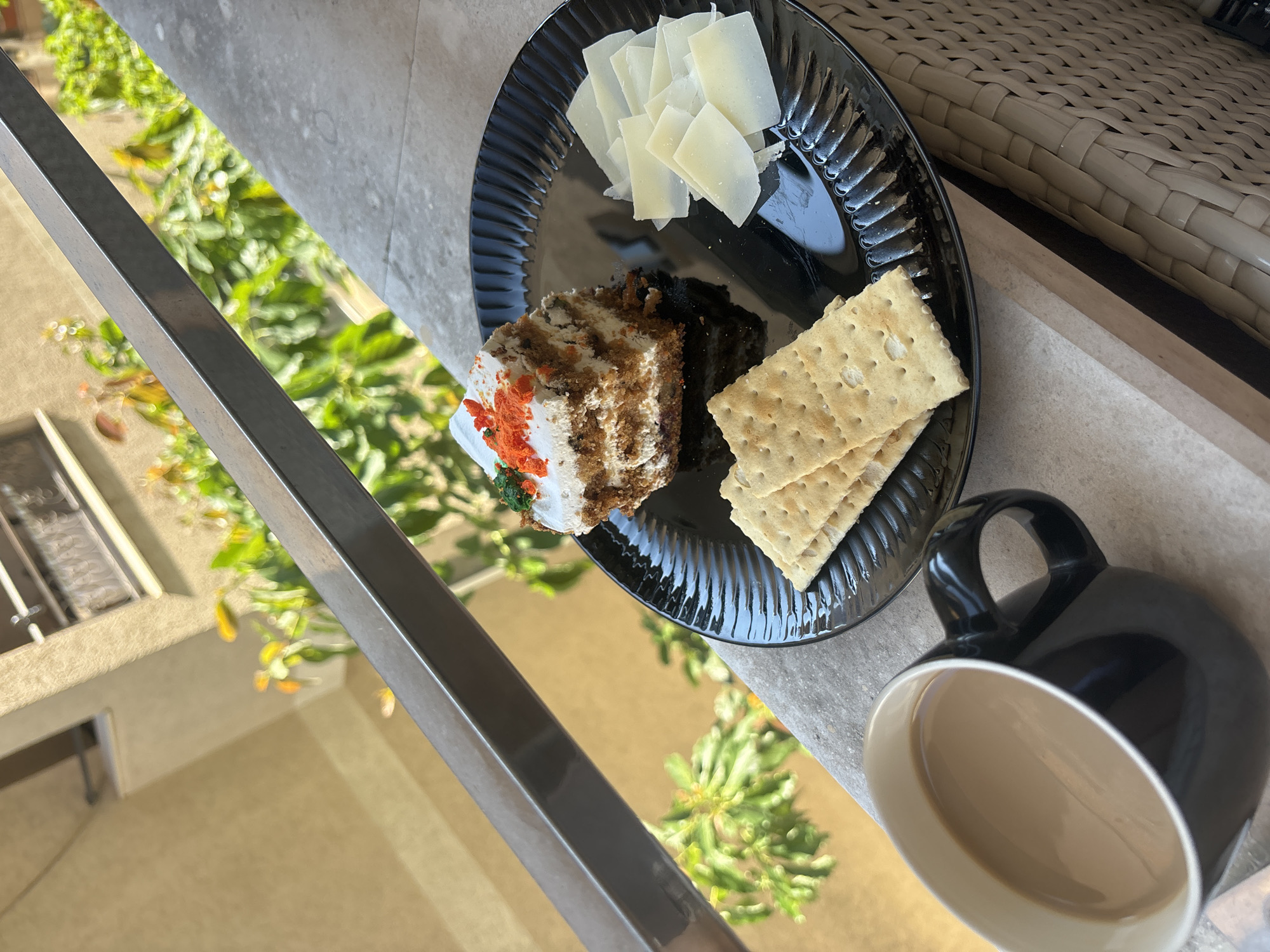 A slice of carrot cake with white frosting and orange sprinkles sits on a black plate alongside several slices of white cheese and two crackers. To the left of the plate is a black mug filled with coffee. The setting appears to be an outdoor balcony or patio, with a metal railing in the foreground and greenery and a building visible in the background. The architectural style of the building and the lush greenery suggest a Mediterranean climate, possibly in Budva, Montenegro, given the context. The scene is illuminated by natural sunlight.