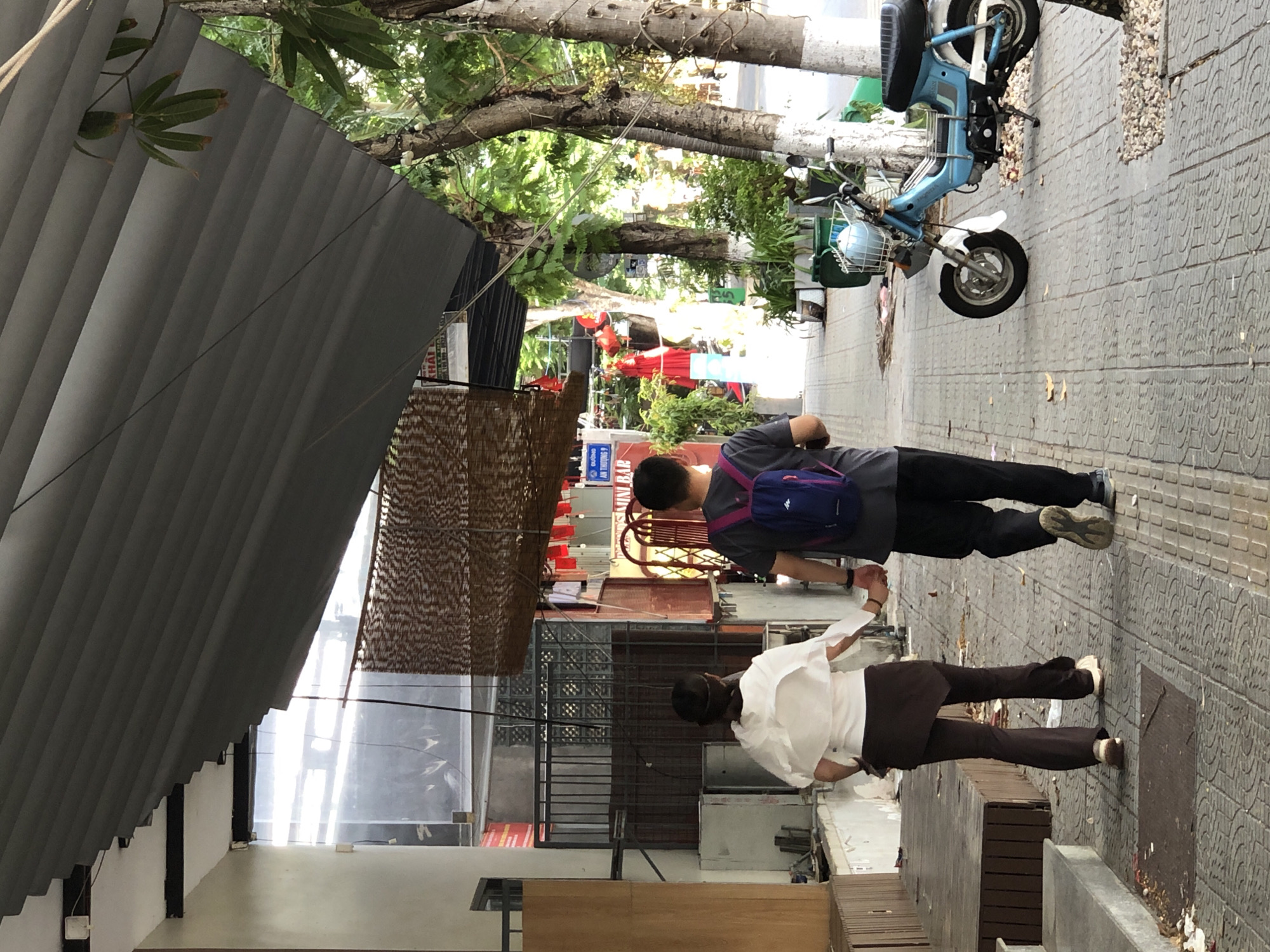 A couple, a man and a woman, walk hand-in-hand down a paved sidewalk in Da Nang, Vietnam, under a gray corrugated metal awning. The woman, in front, wears a white flowy top and brown flared pants. The man, behind her, wears a dark t-shirt with a blue backpack and black pants. A light blue moped is parked on the sidewalk to the right of the couple. The sidewalk is lined with trees and potted plants. The buildings on the left have awnings and windows, and some storefronts are visible. The overall impression is of a warm, sunny day in a busy urban area.