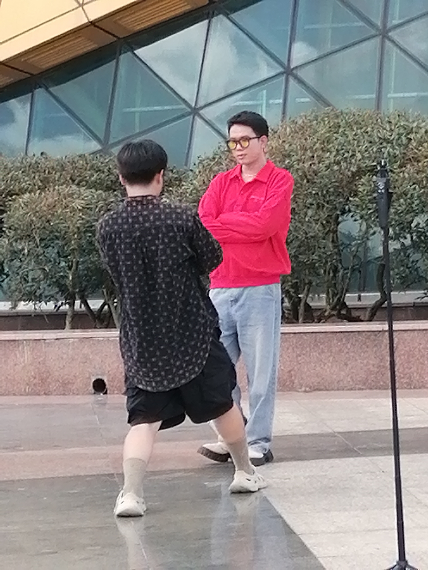 Two individuals are outdoors, positioned in front of a building with a faceted glass facade and a hedge. The person on the left, viewed from behind, wears a dark patterned button-down shirt and black shorts, with white socks and white slip-on shoes. They appear to be in motion, with their left leg extended. The person on the right faces the other, wearing a red long-sleeved polo shirt, light blue jeans, and white shoes. They have yellow-tinted sunglasses and their arms are crossed. A black microphone stand is visible to the right of the frame. The ground is a paved surface with reflective qualities. The setting suggests a public space, possibly a plaza or walkway in Da Lat, Vietnam, given the architectural style and vegetation.