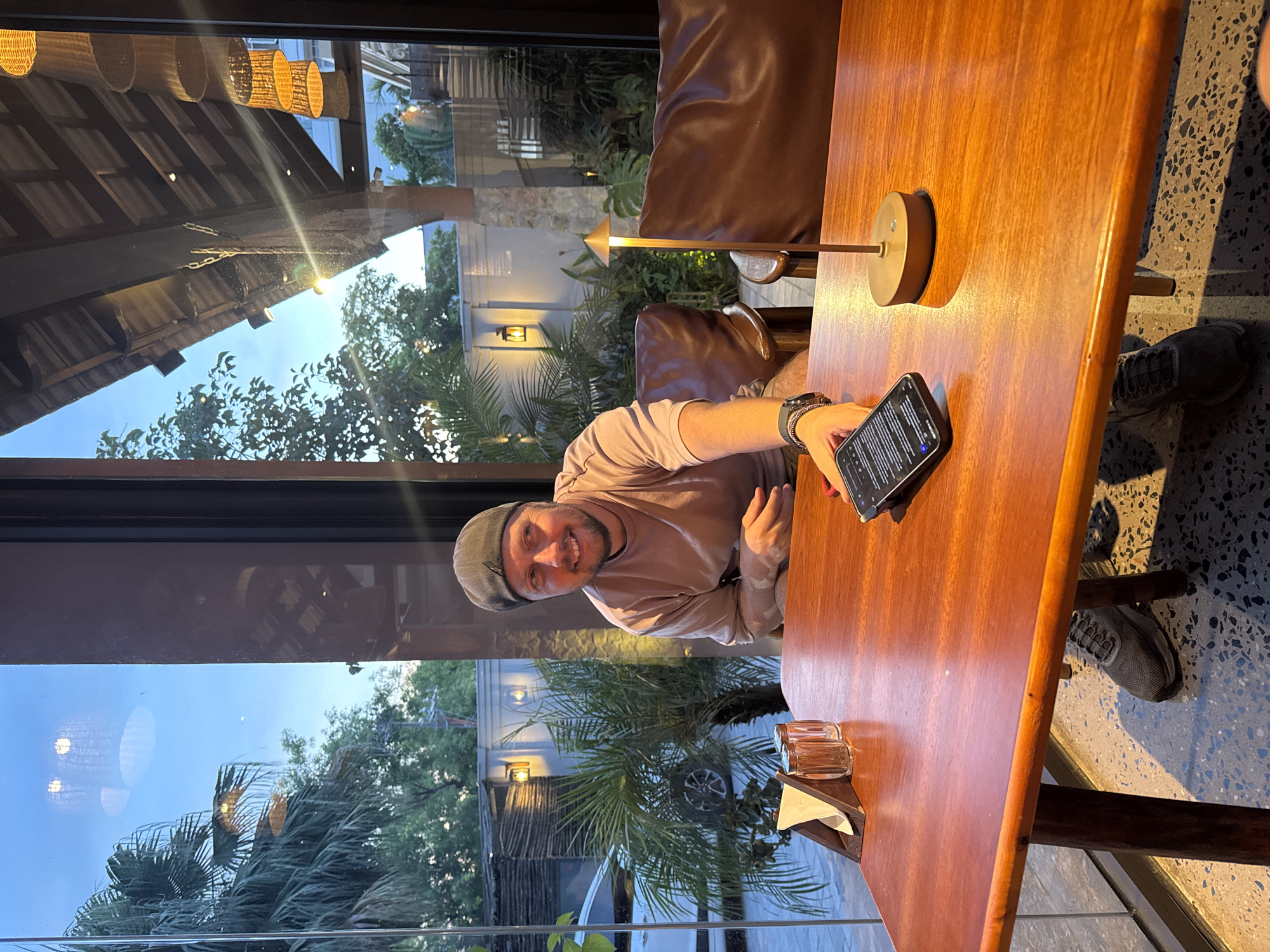 In Nam Hoa Lư Ward, Vietnam, a man wearing a gray cap and a light brown long-sleeved shirt is seated at a wooden table. He is smiling and looking towards the camera, holding a smartphone in his outstretched hand. The table is set with a napkin holder containing napkins and two salt and pepper shakers. A small lamp with a brass base and shade is also on the table. Behind him, large windows offer a view of lush greenery and outdoor lighting. The environment suggests an indoor seating area of a restaurant or cafe with an outdoor patio.