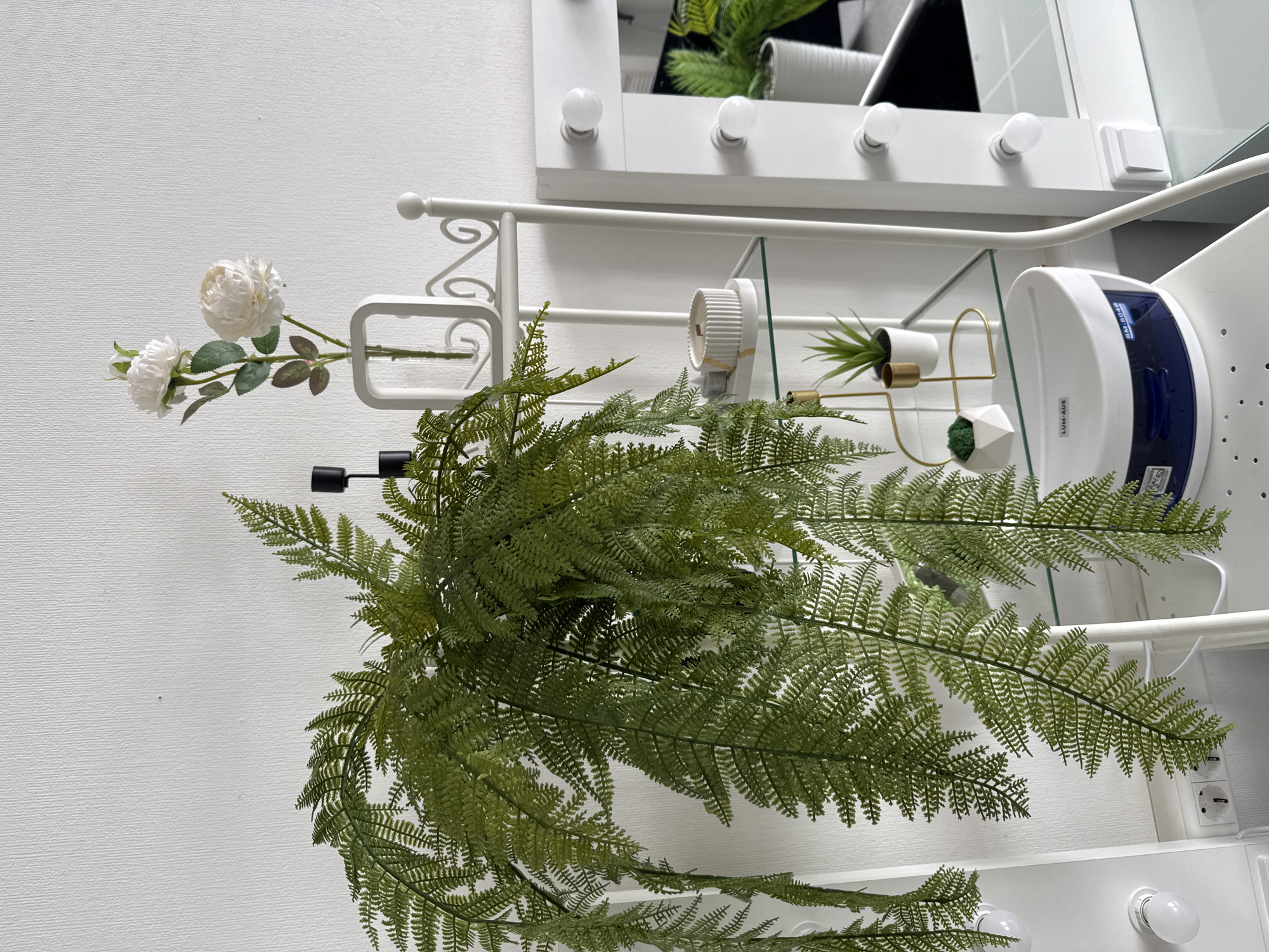The image depicts a still life arrangement on a white shelf against a white wall. A faux fern plant dominates the scene. To the left, there's a faux white rose in a transparent vase with a white frame. Behind the fern, a small potted succulent sits on a decorative gold and white stand. A white electric fan or switch is mounted on the shelf. Further back, a vanity mirror with light bulbs along its frame is visible. A white UV nail lamp is also present. The electrical outlet suggests the scene is indoors.
While the image shows details of interior decor, there are no indicators it's specifically in Chaikovsky, Russia.
