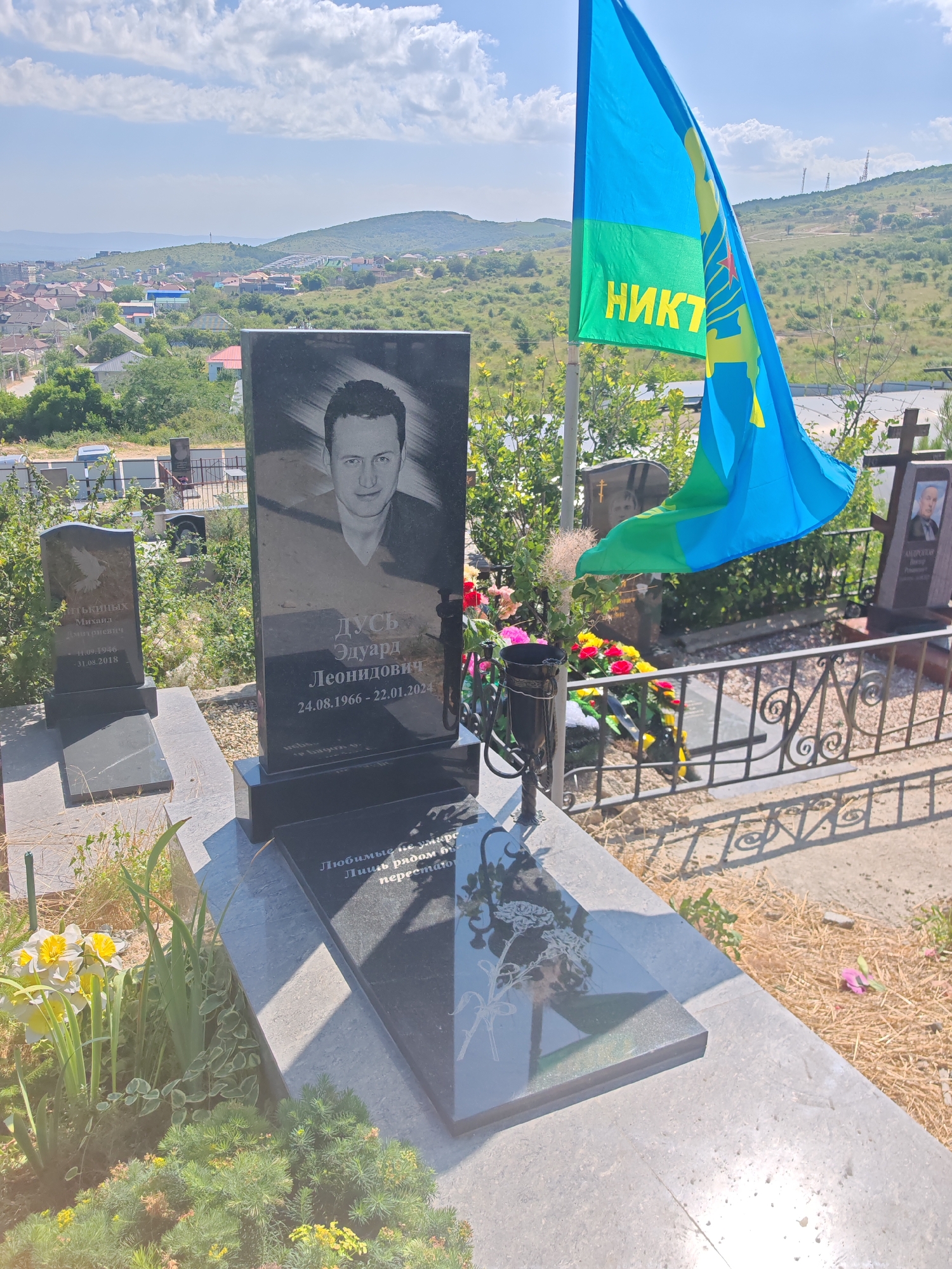 A cemetery in Supsekh, Russia, features a prominent black granite headstone with a portrait of a man and Cyrillic inscriptions. To the right of this headstone, a blue and green flag with yellow lettering and symbols waves from a pole. In the background, a town with buildings and hilly terrain is visible under a partly cloudy sky. Other headstones and grave markers are scattered throughout the scene, with some floral arrangements and greenery present. The ground is covered in dry grass and gravel.