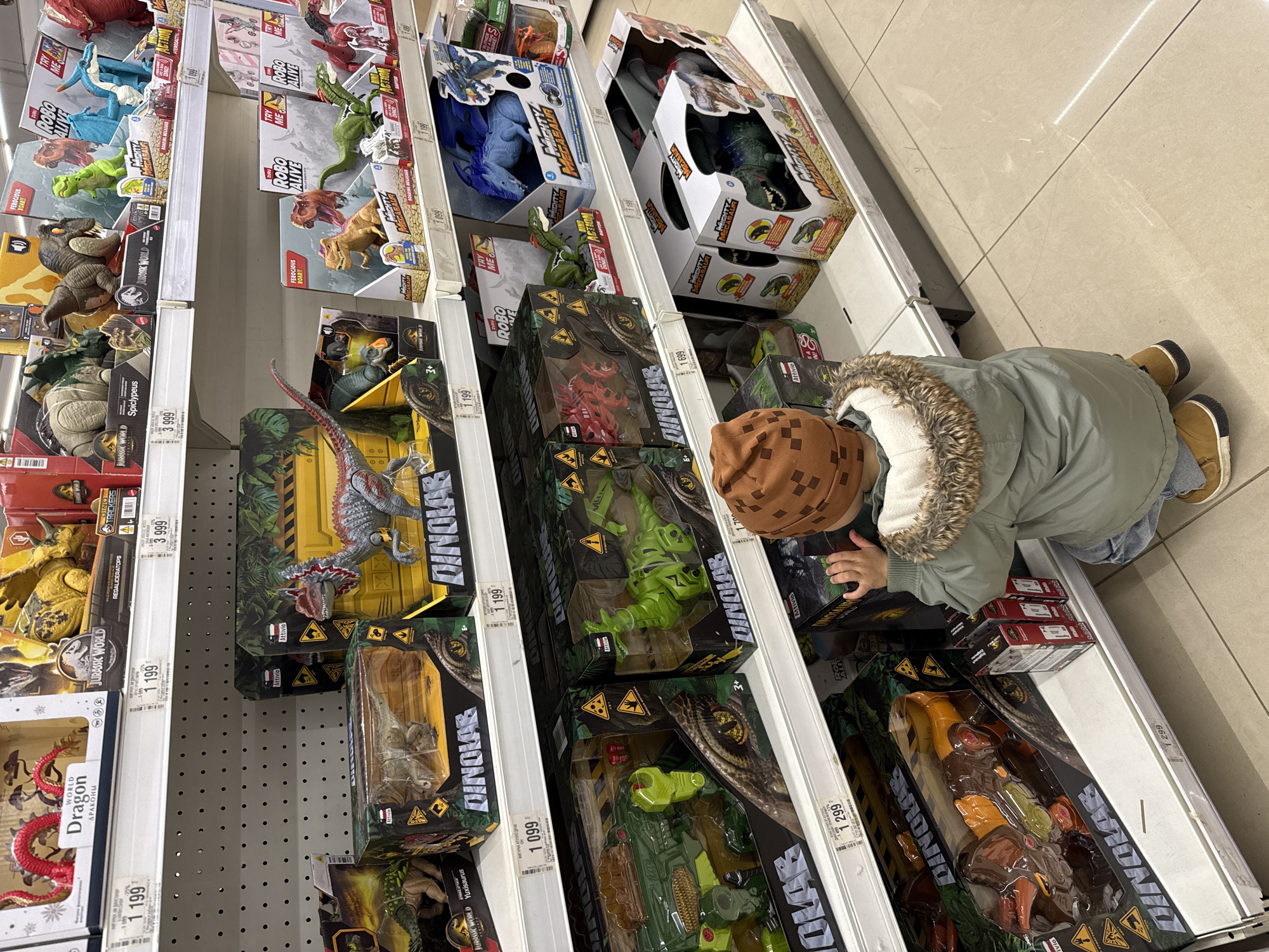 The image shows a child at a store shelf stocked with dinosaur toys, likely in Korobovo, Russia. The child is wearing a green jacket with a fur-lined hood and a brown patterned hat. They are bending down to look at the toys on the lower shelves. The shelves are filled with various dinosaur toys in boxes, mostly from the Jurassic World and Robo Alive brands. The prices of the toys are visible on the shelf tags, ranging from 1,099 to 3,999. The floor is tiled with light-colored square tiles.