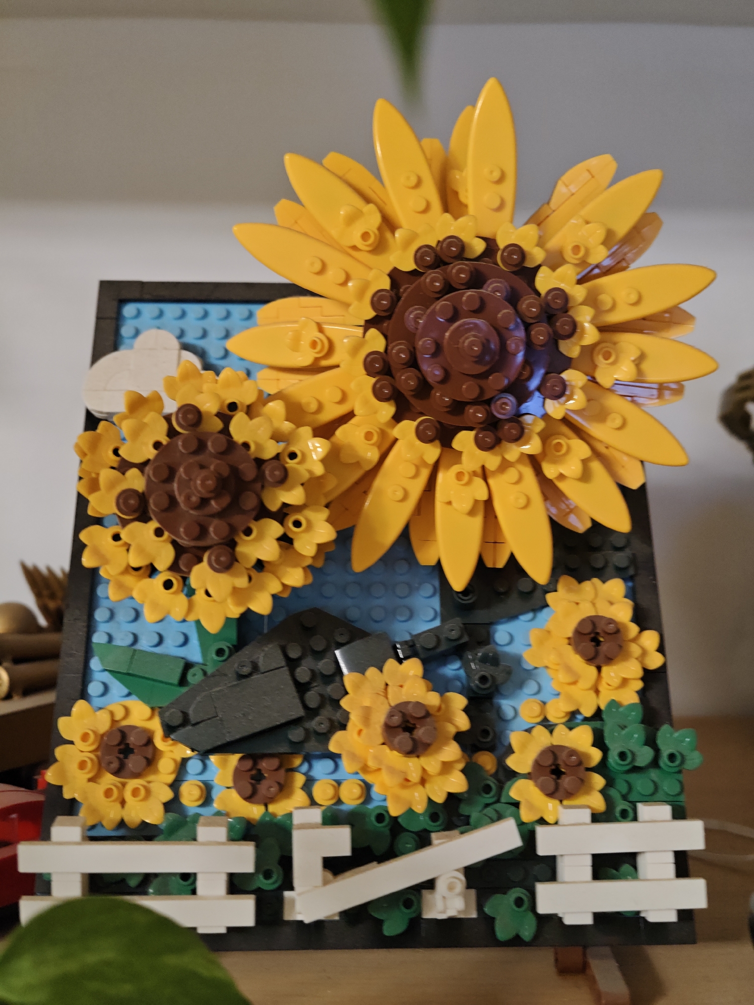 The image displays a close-up, eye-level, static shot of a LEGO diorama. The diorama depicts a garden scene with several sunflowers, a white picket fence, and a sky with clouds, all constructed from LEGO bricks. The largest sunflower, with bright yellow petals and a brown center, dominates the upper portion of the frame. Smaller sunflowers are arranged in the foreground and midground, interspersed with green LEGO leaves. The background features blue LEGO bricks representing the sky, with white LEGO cloud elements. A dark-colored LEGO structure, possibly representing garden tools or a planter box, is situated in the midground. The diorama is placed on a wooden surface, and the background above the diorama appears to be a plain, light-colored wall. The lighting is soft, with no harsh shadows. There are no people visible in the image. The scene is stationary, with no action taking place. The diorama itself appears to be a decorative item, possibly a display piece. The location is not specified within the image or discernible from its content.