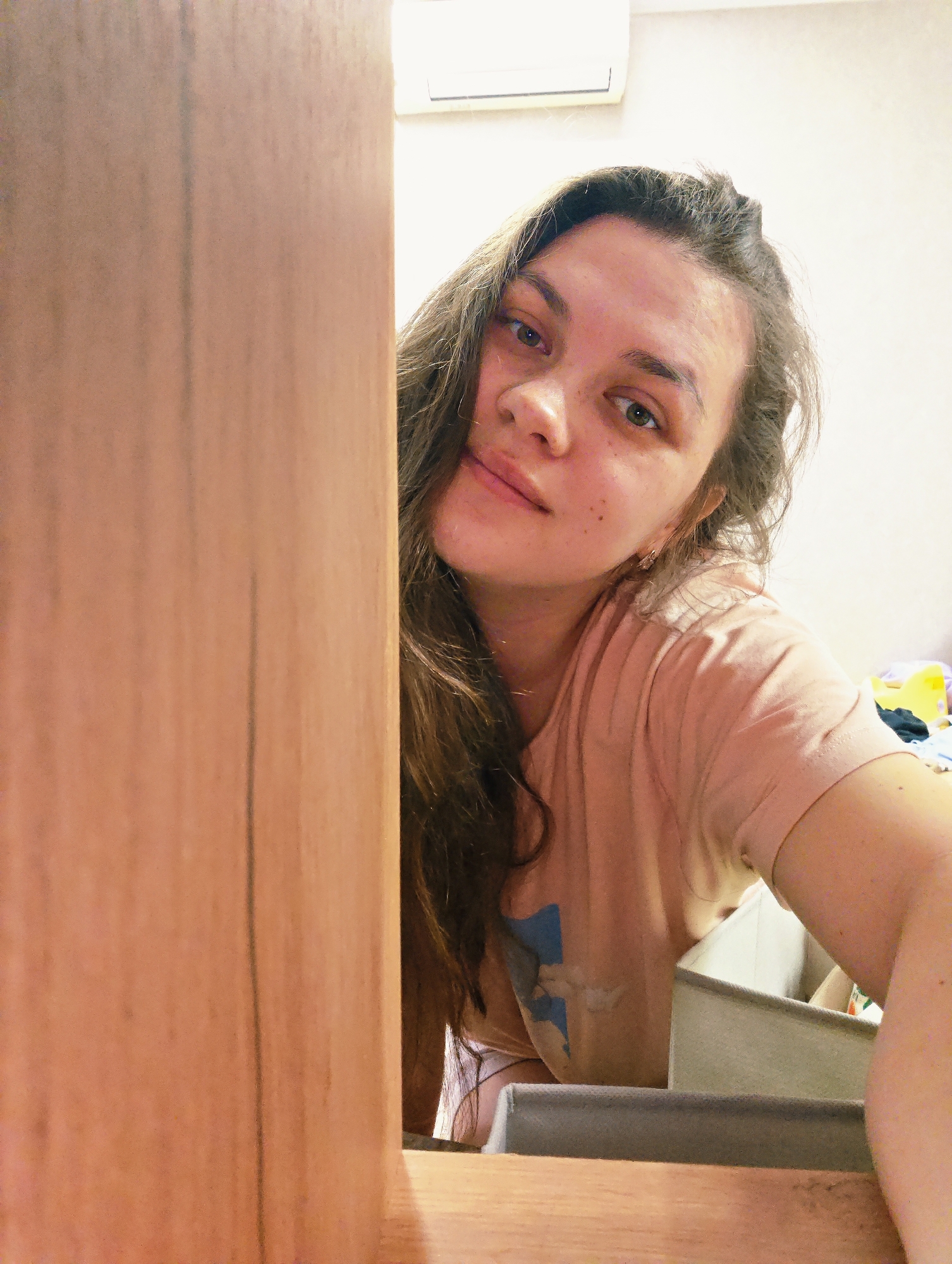 A young woman with long brown hair and fair skin is partially visible behind a light brown wooden structure. She is wearing a pink t-shirt and appears to be looking at the camera. Behind her, there is a white wall and a white air conditioning unit. A grey fabric container is also visible. The location is Chaikovsky, Russia.