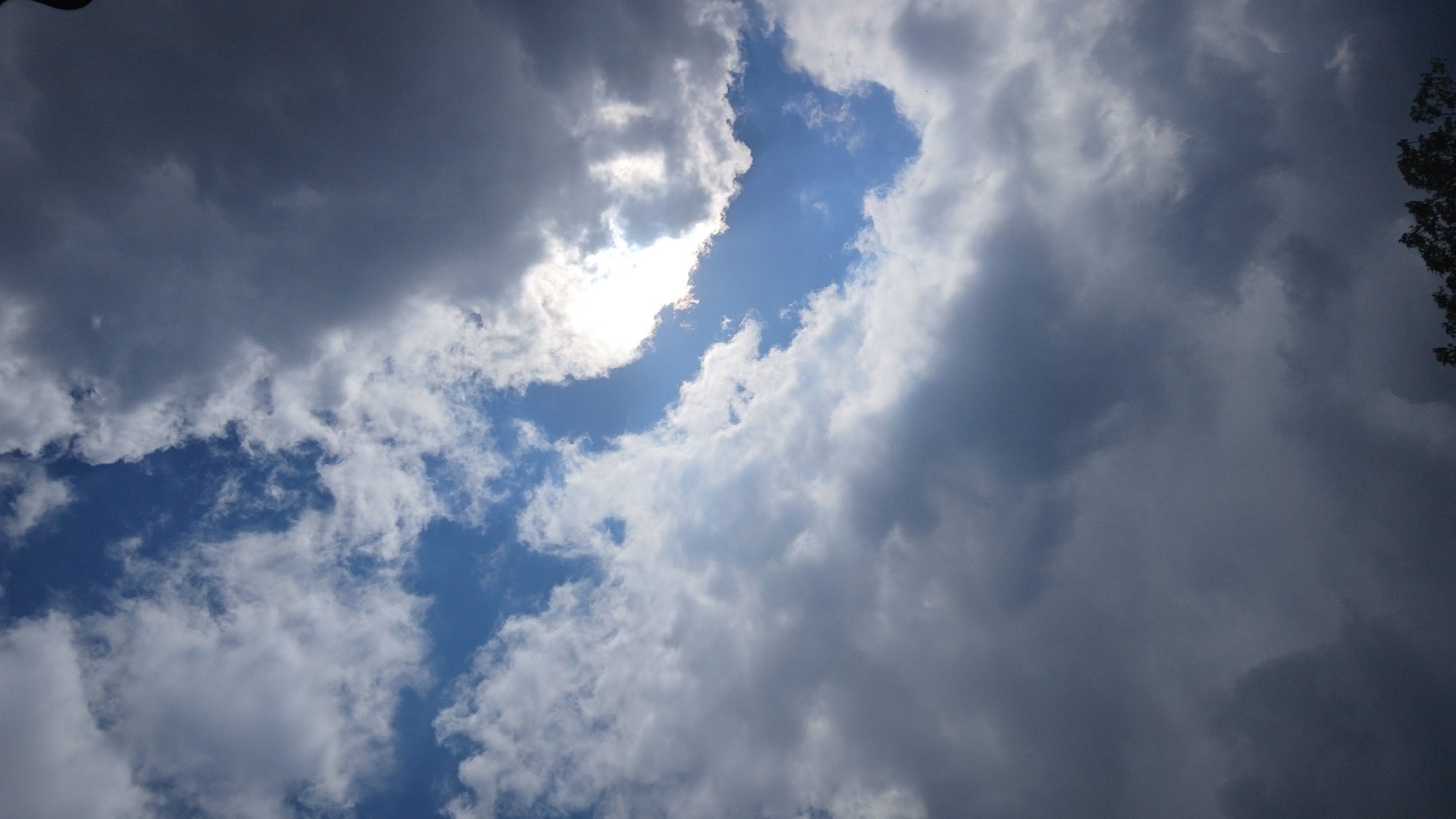 The image shows a sky filled with clouds, with patches of blue visible. The sun is partially obscured by clouds, creating a bright spot in the upper center. The clouds range in color from bright white to dark gray. Some foliage is visible in the upper right corner. The location is Unknown, Unknown.