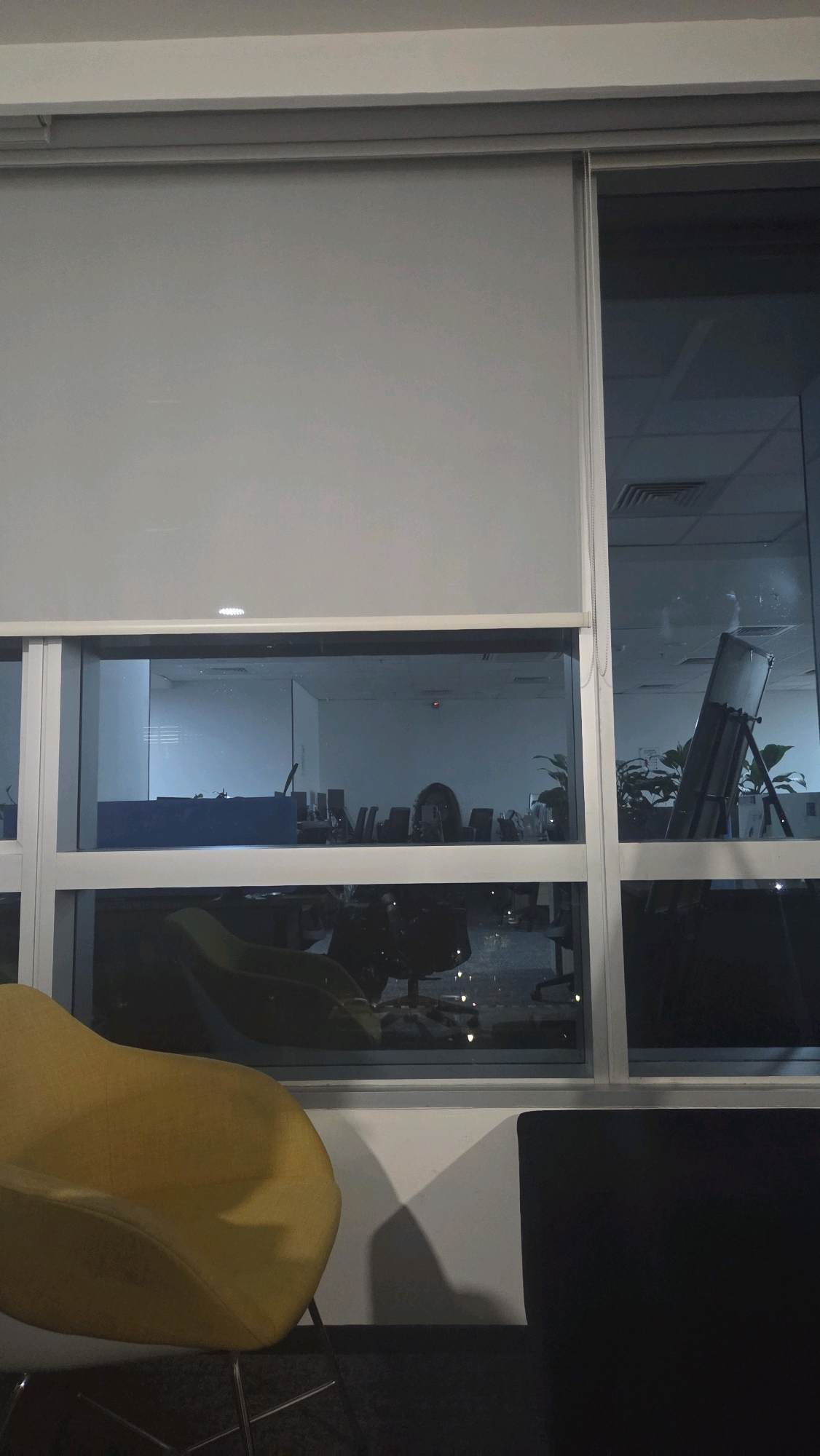 The image shows an interior view of an office space in Bengaluru, India, taken at night. A large window spans across the scene, partially covered by a light gray roller blind. Through the window, an office environment is visible, with rows of desks and office chairs. In the middle section of the window, a person's reflection or presence is discernible, appearing as a dark silhouette with visible hair. Plants are also present within the office. To the right of the window, a projector screen on a stand is visible. In the foreground, a bright yellow armchair with metal legs occupies the left side of the frame. A dark sofa or chair is partially visible on the right. The lighting suggests it is evening or nighttime, with some artificial lights visible within the office.