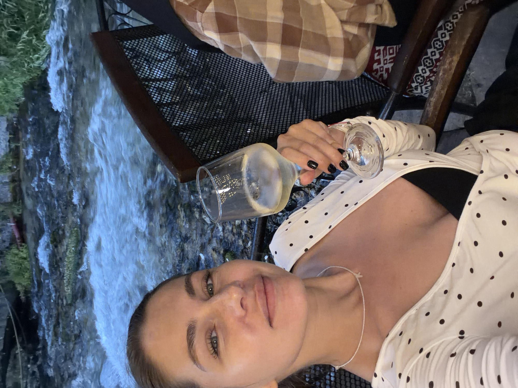 The media file shows a woman taking a selfie in an outdoor setting. She is holding a wine glass filled with a white beverage and wearing a white polka-dot top with a black undershirt. Her nails are painted black. The background features a stream of water flowing over rocks, with some greenery to the left. To the right, there's a glimpse of a table and chairs, suggesting a dining or seating area, possibly at a restaurant or cafe. A checkered blanket is visible on one of the chairs. The lighting suggests it might be evening or a shaded area. The overall mood appears relaxed and leisurely. The woman's facial expression is soft and pleasant. No visible text is present in the image. The location context provided is Travnik, Bosnia and Herzegovina.