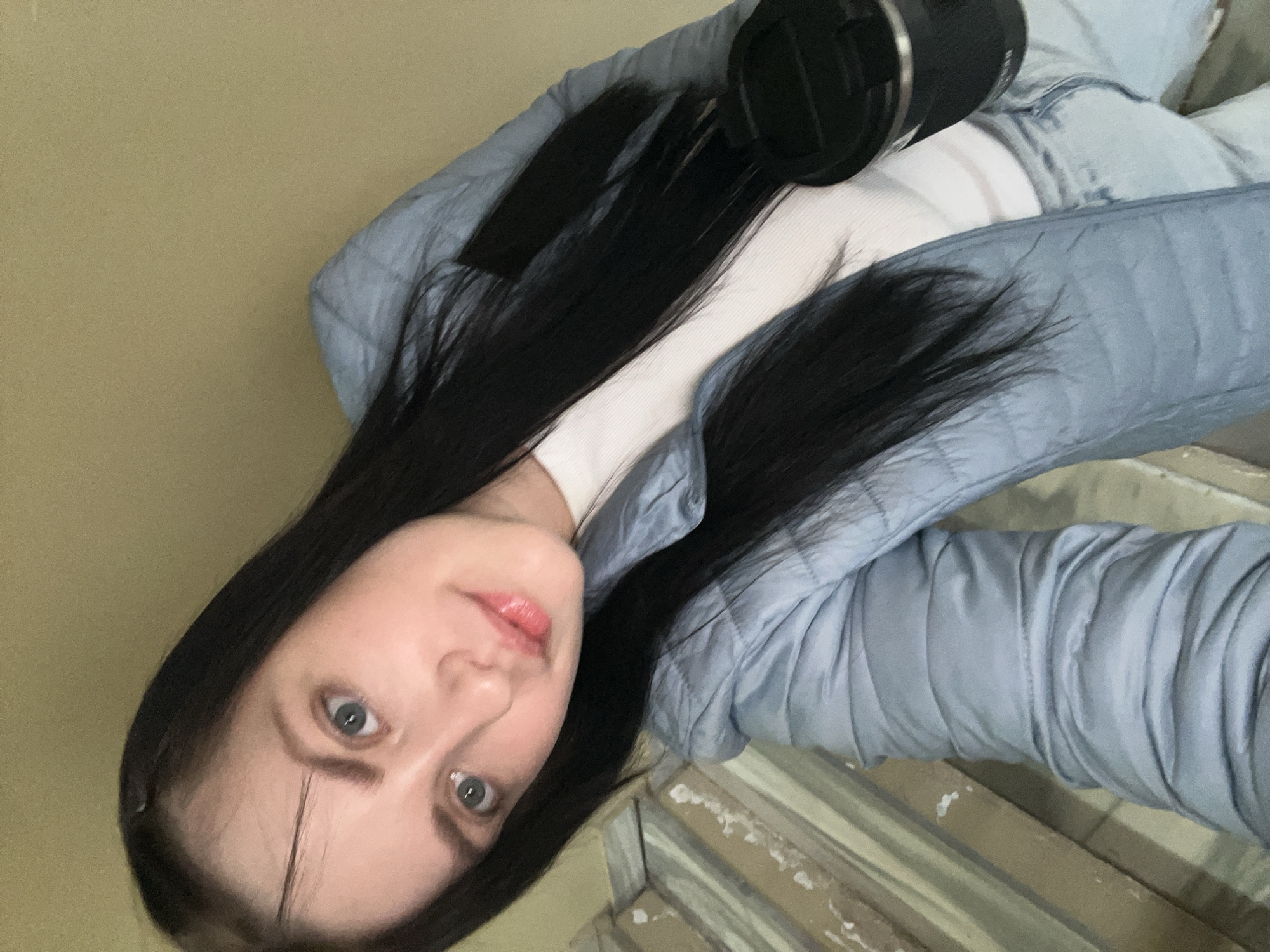 A woman with long, dark hair, blue eyes, and light skin is posing for a photograph. She is wearing a white top and a light blue quilted jacket. A black camera lens is positioned near her upper chest. The background includes a yellow-toned wall and a set of wooden beams or slats. The image was taken in Марковский, Russia.