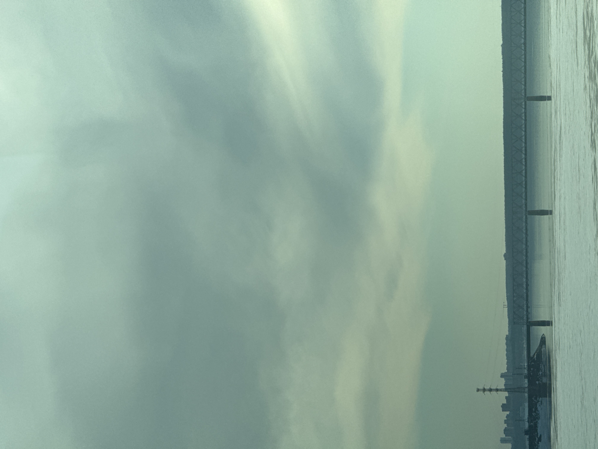 The image depicts a bridge spanning water, with the city of Пальники, Russia visible in the background. The sky is overcast, creating a muted, hazy atmosphere. The bridge structure is primarily metal and appears to be a truss bridge. Vertical supports are visible at regular intervals along the bridge's length, and a few buildings and structures are visible along the shoreline. The water appears calm, with some indication of waves or ripples.