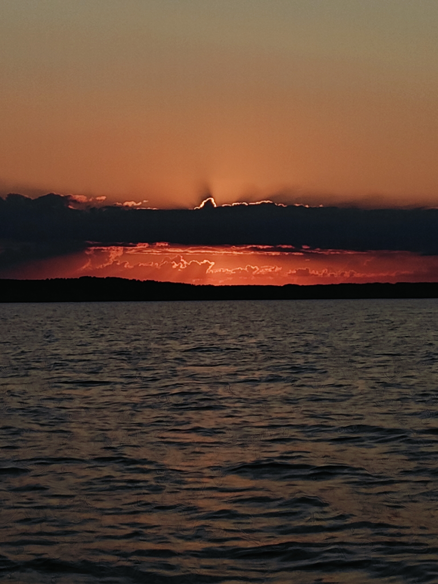 This is a vertical, medium-shot, photograph of a sunset over a body of water. The sky is filled with shades of orange and yellow, with dark, heavy clouds obscuring the sun. The sun is partially visible behind the clouds, casting rays of light that create a halo effect. A dark, silhouetted landmass with a treeline is visible on the horizon, separating the sky from the water. The water in the foreground is dark and rippled, reflecting the colors of the sky. The scene is devoid of any people or man-made structures, creating a sense of natural beauty and solitude. The location is identified as Chaykovsky, Russia.